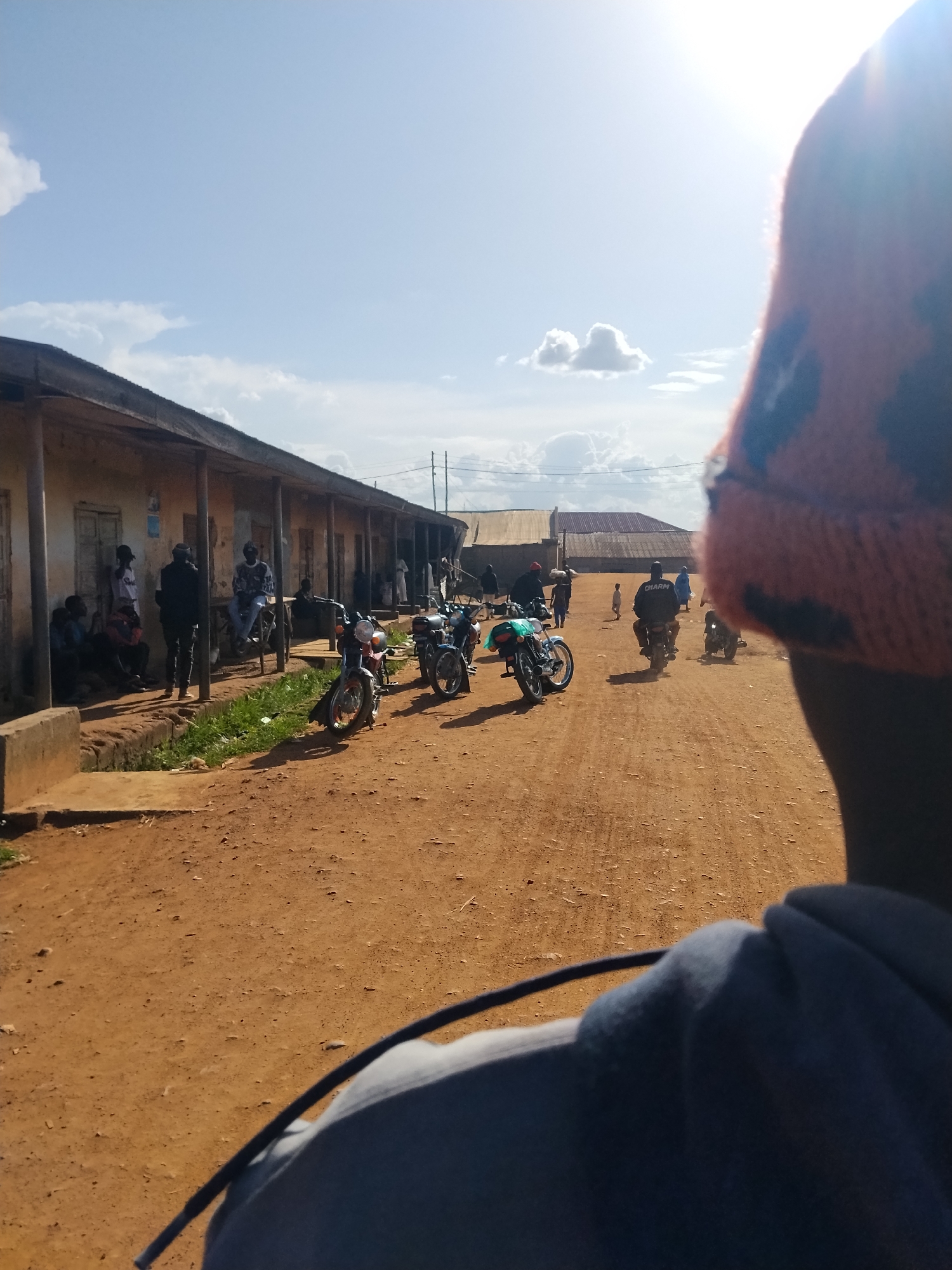The image depicts a sunny day in Mangu Town, Nigeria, with a clear blue sky dotted with fluffy white clouds. The scene is a dusty, unpaved street lined with simple, one-story buildings. The buildings appear to be made of mud or concrete with corrugated metal roofs.
Several people are present, gathered in small groups along the street. Some are standing near the buildings, while others are sitting. A few motorcycles are parked along the side of the street, and some people are seen riding motorcycles in the distance. The presence of motorcycles suggests that they are a common mode of transportation in this location. The overall atmosphere appears to be casual and community-oriented, with people socializing and going about their daily activities.
The time of day is likely mid-morning or early afternoon, given the bright sunlight and the shadows cast on the ground. The weather is warm and sunny, typical for a tropical climate. The unpaved street, simple architecture, and the presence of motorcycles are strong indicators of a rural or semi-urban setting in Nigeria.
In the foreground, the back of a person wearing a blue hooded sweatshirt and an orange beanie is visible, suggesting the photo was taken from the perspective of someone riding on the back of a motorcycle or walking behind someone.
No visible text is discernible in the image.