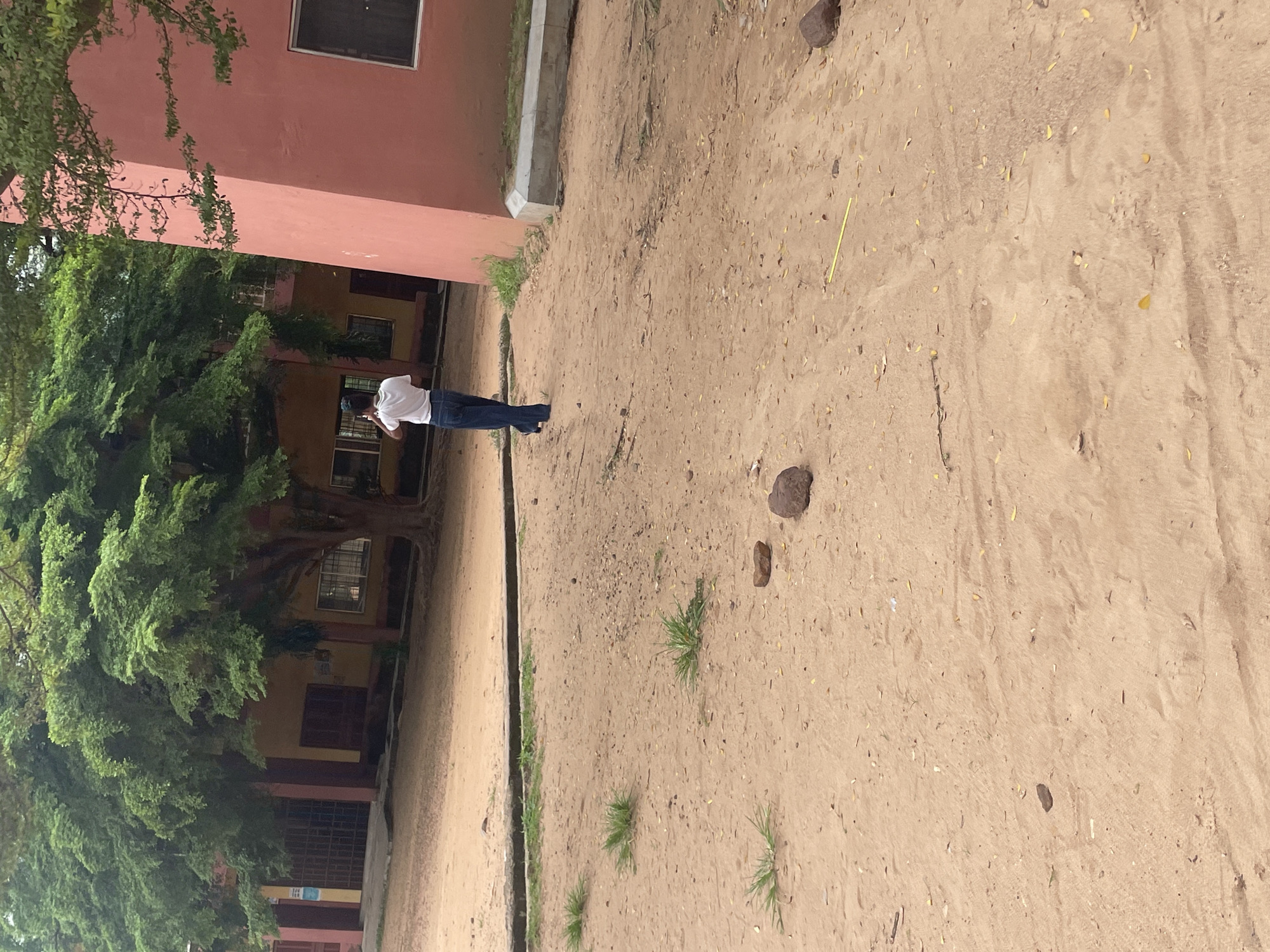 A young person wearing a white t-shirt and blue jeans walks away from the camera on a sandy, dirt path in front of a pink, modern-style building. Large trees with dense green foliage are visible on the left side of the image, partially obscuring a tan building with red accents and barred windows. The ground is covered in sand, with scattered leaves, small rocks, and patches of grass. In the background, the pink building has a large, dark-paned window and a concrete curb. The scene appears to be an outdoor area, possibly a school or public space, in Lokoja, Nigeria, given the distinctive pink building architecture and abundant vegetation often found in the region.
