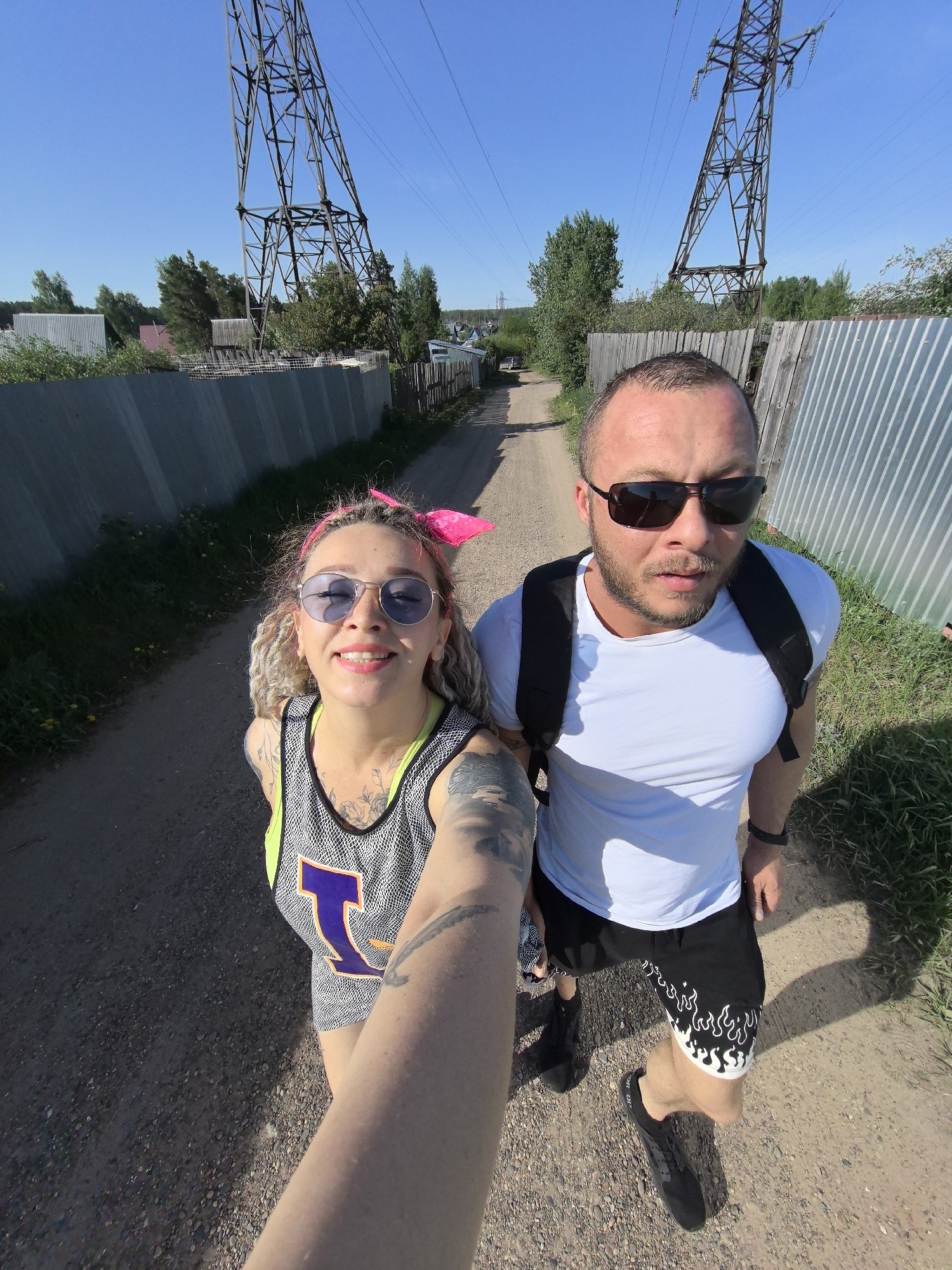 A selfie taken by a woman with blond dreadlocks, wearing purple sunglasses and a neon yellow-trimmed tank top with a large purple "L" on it, showing tattoos on her arm and chest. Beside her is a man with sunglasses, wearing a white t-shirt and black shorts with white flame designs, with a black backpack on. They are standing on a dirt road between metal fences and greenery. Overhead are large metal utility towers and power lines. The location is Noviy, Russia.