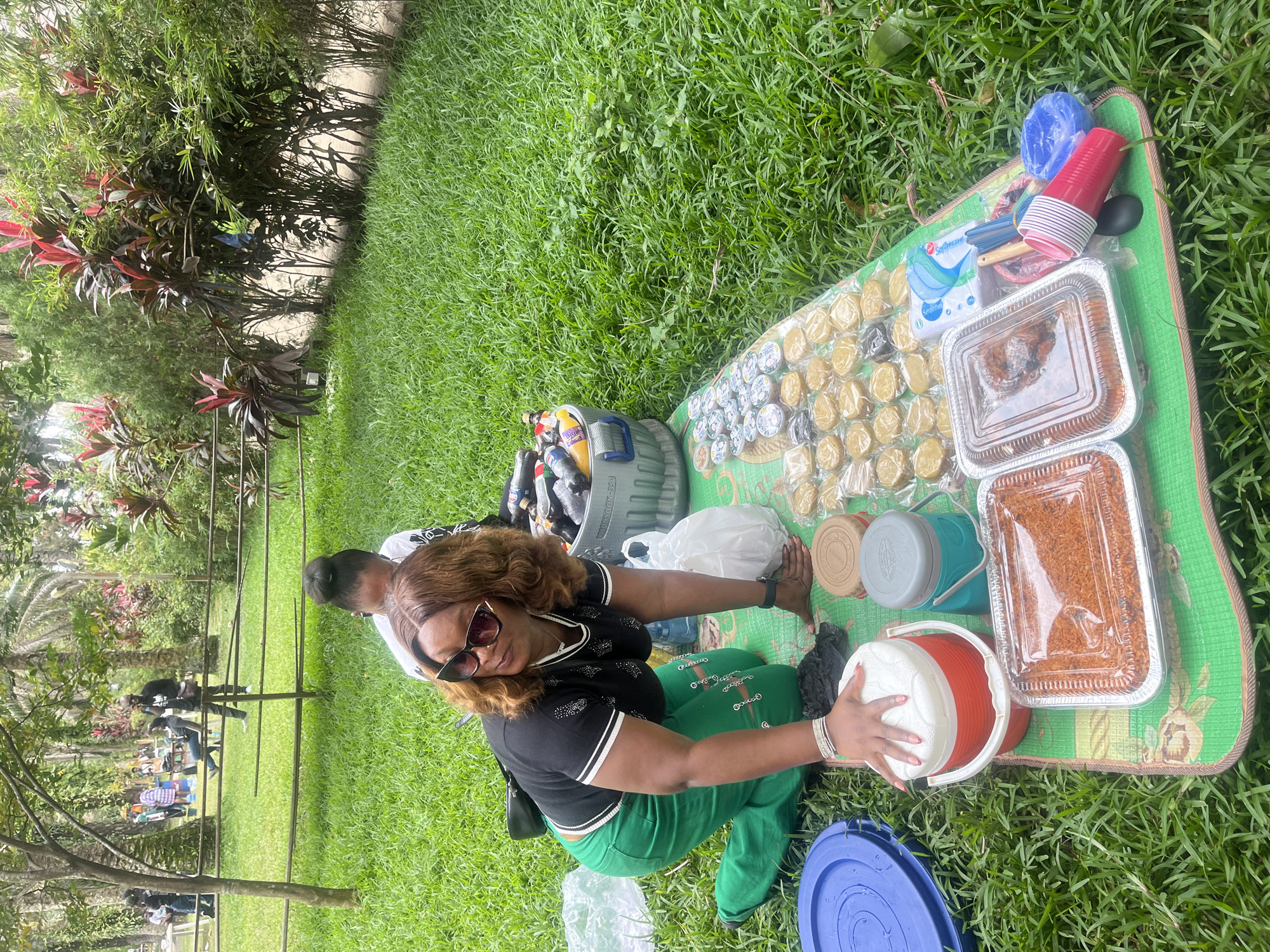In Lekki, Nigeria, a woman is kneeling on a picnic mat in a grassy area, preparing food. She is wearing a black shirt, green pants, and sunglasses. A cooler and various food containers are spread out on the mat. In the background, other people are visible, some sitting and others standing, in a park-like setting with trees and foliage. The scene suggests a casual outdoor gathering.
