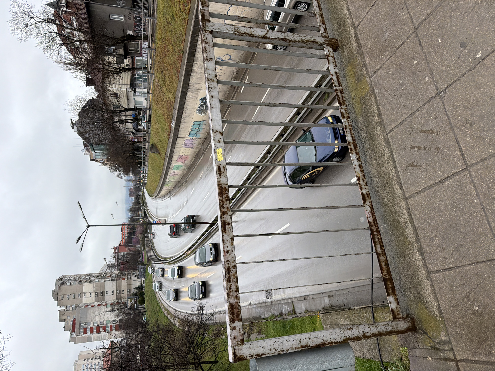 This image captures an elevated view of a multi-lane urban road in Sofia, Bulgaria, under an overcast sky.
In the immediate foreground, a heavily rusted metal railing of a pedestrian overpass extends across the lower portion of the frame, partially obscuring the view of the roadway below. The concrete tiled surface of the overpass platform is visible in the lower right, displaying wet patches and some yellowish staining near the railing.
Below the overpass, a wet, divided highway/boulevard stretches into the distance, with vehicles traveling in both directions. The road surface is visibly reflective from recent rain. In the rightmost lane of the carriageway moving toward the viewer, a dark blue Subaru Impreza sedan with a yellow graphic on its side is prominent, displaying a Bulgarian "CB" series license plate. Behind it, a silver SUV is partially visible. Further back in the same direction, a dark grey SUV and a black sedan are discernible. In the opposing carriageway, several other vehicles, including dark grey, white, and silver cars, are visible with their headlights illuminated. White lane markings are distinct on the wet asphalt.
Concrete retaining walls flank both sides of the road, with the wall on the right (from the viewer's perspective) featuring extensive multi-colored graffiti. Grassy embankments rise from behind these walls. Street lighting poles are spaced along the road, some with illuminated lights.
The background on the left features a large, multi-story residential building with a grey and white facade, accented with red panels, alongside other high-rise structures. Bare deciduous trees line the roadside. On the right, a row of older, mixed-use buildings with varying architectural styles and pitched roofs is visible, also bordered by bare trees. Distant hills or mountains are faintly discernible through the hazy, overcast conditions in the far background. No identifiable individuals are visible.
