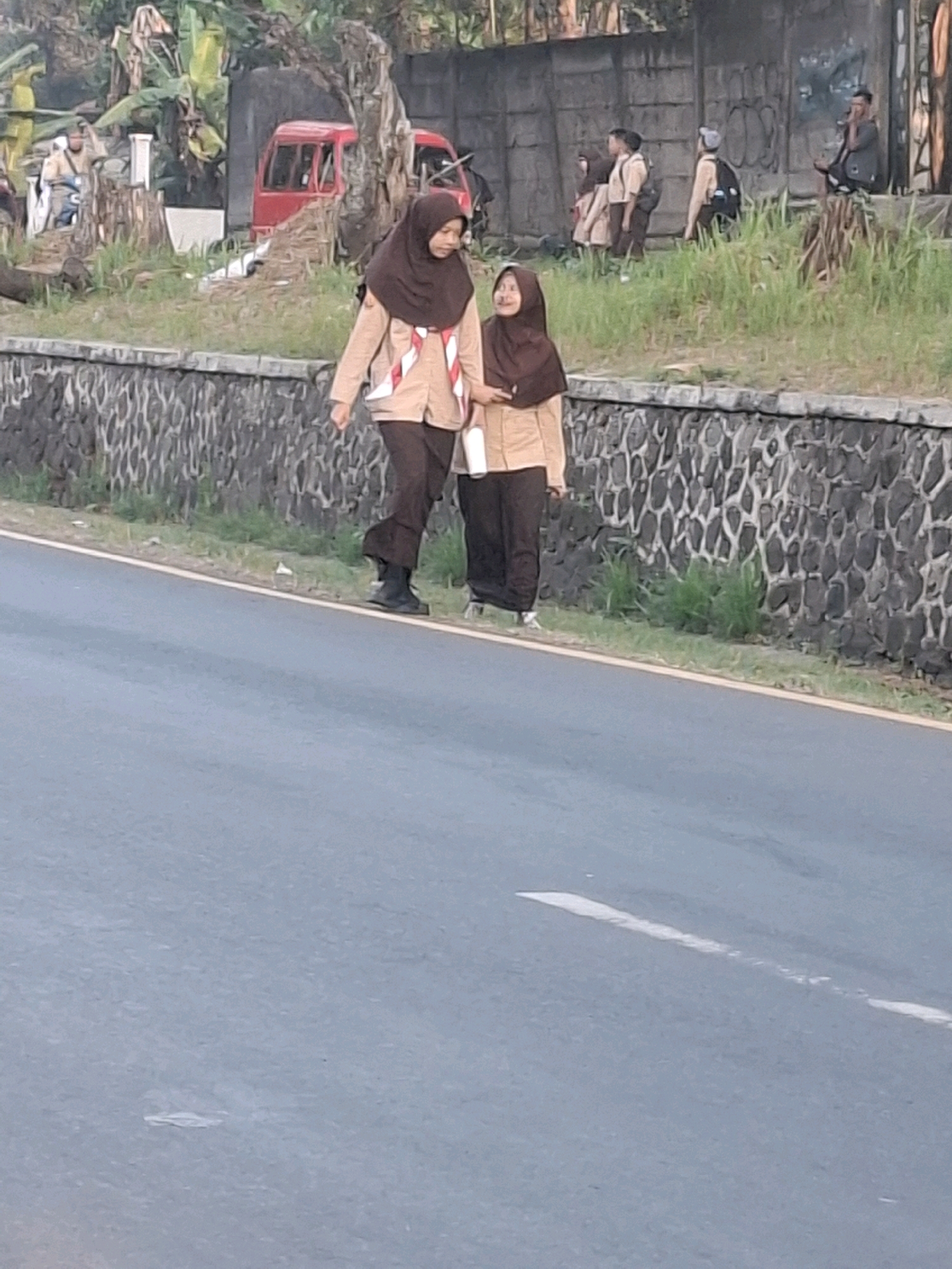 Two young individuals wearing scout uniforms, one older and one younger, walk along a paved road next to a stone wall. The older individual is on the left, wearing a tan long-sleeved shirt, dark pants, black boots, and a brown hijab. The younger individual is on the right, wearing a similar tan uniform with dark pants and a brown hijab. They are walking together, appearing to be engaged in conversation. The background shows a grassy area, trees, and a red vehicle parked behind a fence. The environment suggests an outdoor setting in what appears to be a rural or semi-urban area of Sirnagalih, Indonesia.