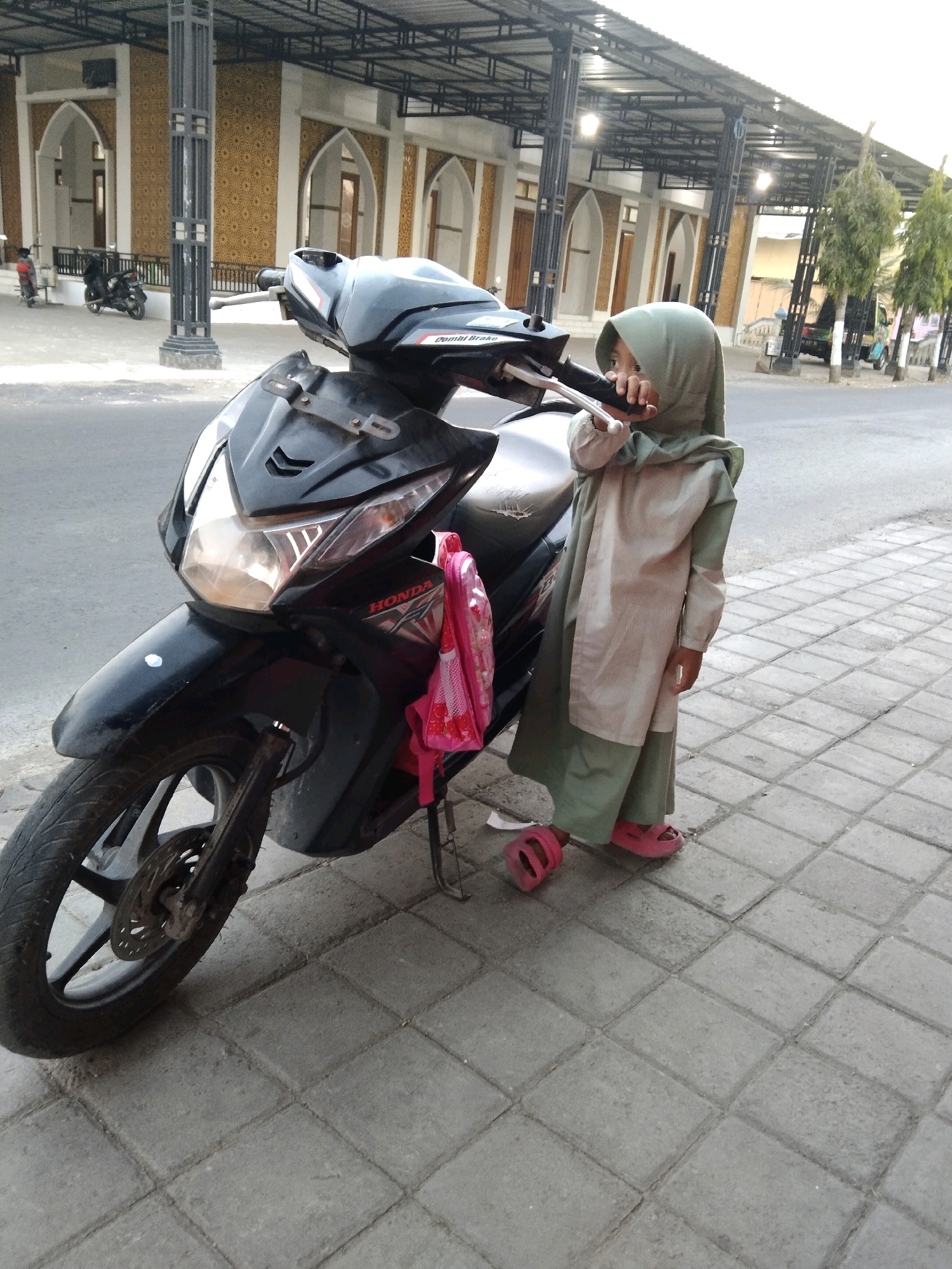 A young girl wearing a light green hijab and a light brown and green robe stands next to a black motorcycle on a paved sidewalk. She is holding the handlebars of the motorcycle and appears to be looking at something to her right. She is wearing pink sandals. A pink backpack with a floral pattern is hanging on the side of the motorcycle. The motorcycle has the Honda logo visible on its front. In the background, a building with arched doorways and decorative patterned walls is visible. The building appears to be a mosque. Several trees are also present. The scene is set in Glagahan, Indonesia, as indicated by the context.