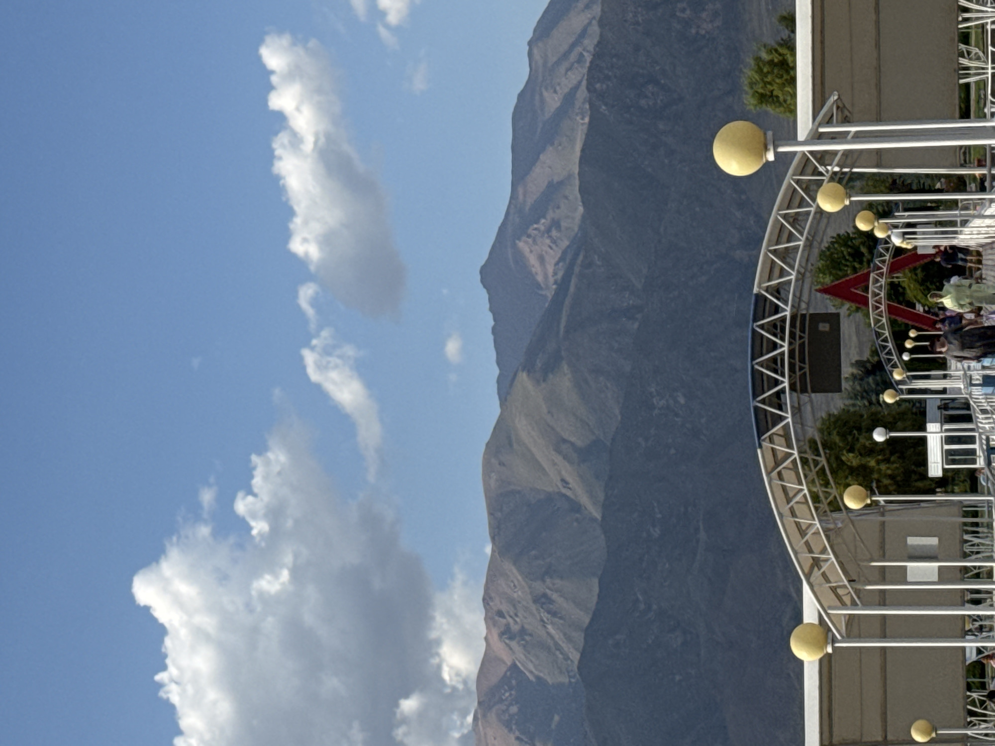 An eye-level, medium shot captures a street scene in Sary-Oy, Kyrgyzstan, under a clear blue sky dotted with white clouds. In the foreground, a white, arched canopy structure spans the width of the image, supported by white poles. Several yellow spherical lights on poles are visible along the canopy, along with some white, round lights. People are seen walking under the canopy, dressed in casual attire, including one person in a light green hooded outfit. In the mid-ground, several trees are visible behind the canopy. A large, red, angular structure, possibly a sign or part of a building, is partially visible behind the canopy. In the background, imposing, rugged, and barren mountains dominate the landscape, their slopes showing signs of erosion and various geological formations. The overall impression is a sunny day in a semi-urban area with a dramatic mountain backdrop.