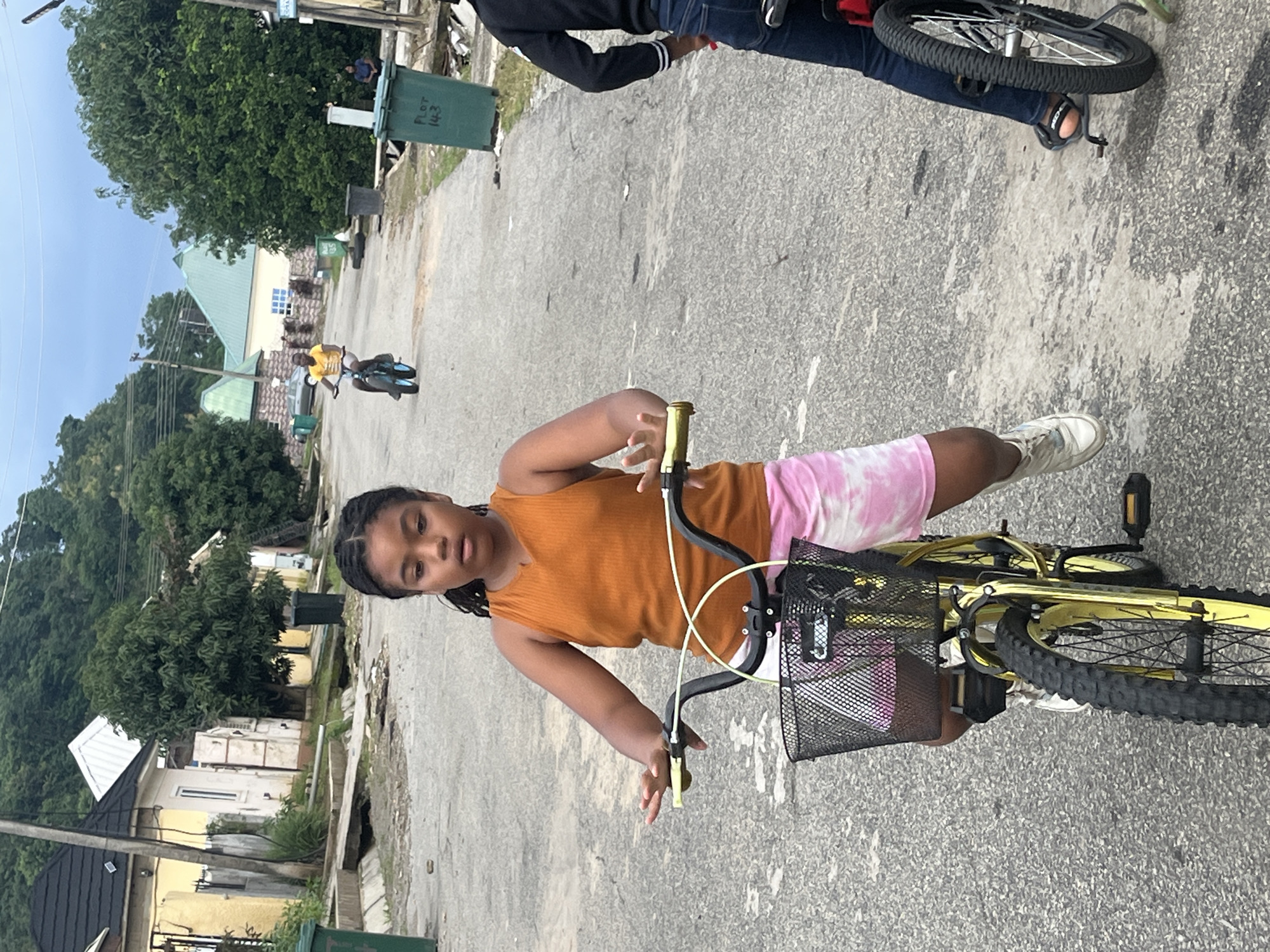 A young girl with braided hair and an orange tank top is riding a yellow bicycle with a black basket in the foreground of the image. She is wearing pink tie-dye shorts and white sneakers. In the background, another child is riding a blue bicycle, and a person in a black jacket and blue jeans is also on a bicycle. The scene takes place on a paved street in what appears to be a residential area in Abuja, Nigeria, with houses and trees lining the road. Trash bins are visible on the sides of the street. The sky is overcast, suggesting it might be early morning or late afternoon.