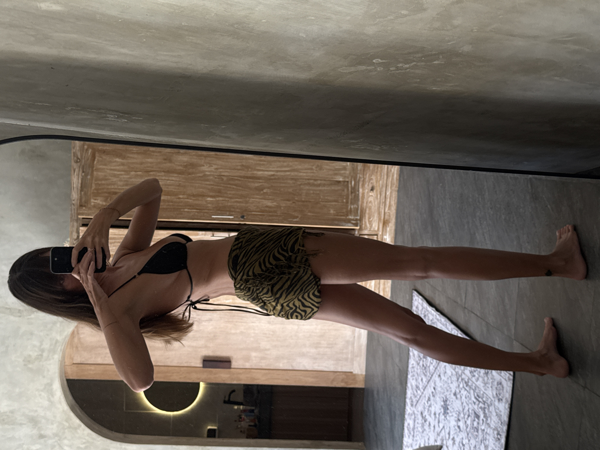 A woman in a black bikini top and zebra-print shorts is standing in front of a mirror, taking a picture of herself with her cell phone. The woman has brown hair and is wearing a bracelet. She is inside a bathroom. In the background, there is a wooden cabinet and a decorative light fixture. The floor is gray. Based on architectural styles and decor, the location is likely North Kuta, Indonesia.