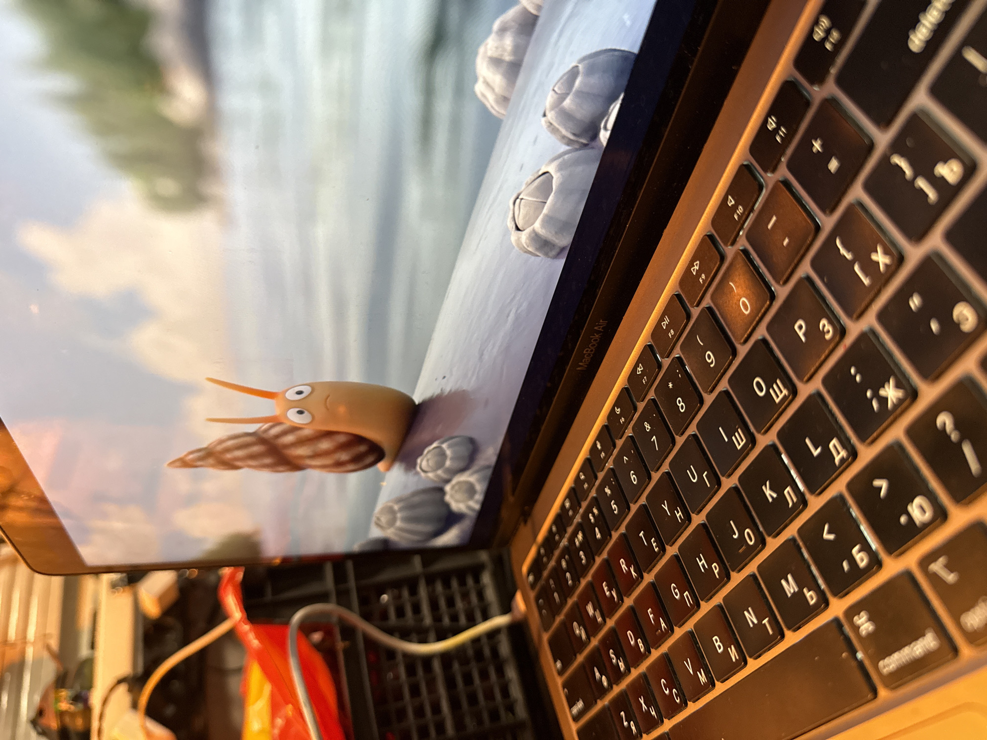The image shows a close-up of a laptop, specifically its keyboard and a portion of the screen. The screen displays a cartoonish orange snail with large eyes and a spiral shell, sitting on a surface next to some barnacle-like objects. The background on the screen appears to be a body of water with trees and a cloudy sky. The laptop is open, and the keyboard is visible, showing keys with Cyrillic characters. To the left of the laptop, part of a red object and a black grate structure are visible, suggesting an indoor setting, possibly a workshop or a room with equipment. There is no indication of the city of Козет, Russia in the image. The lighting suggests it is either daytime or artificial lighting is being used.