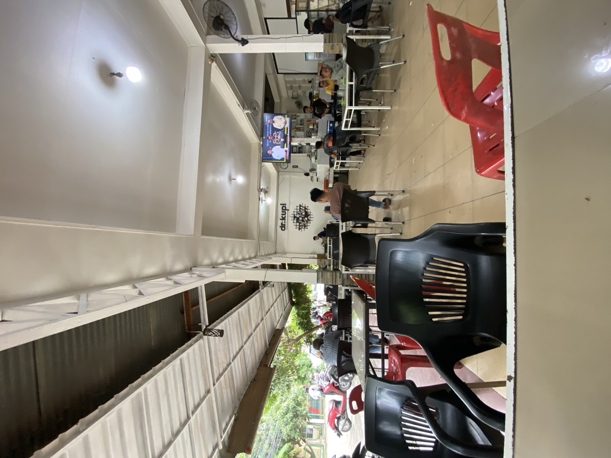 This media file captures a casual indoor/outdoor cafe or restaurant scene, likely a "kopi" shop, in Lhokseumawe, Indonesia. The setting appears semi-open, with an interior dining area connected to an exterior covered space.
Numerous people, predominantly men, are present, engaged in relaxed activities. Some are sitting at tables, watching a flat-screen television mounted on the wall, which displays what appears to be a news broadcast or talk show featuring two individuals. Other patrons are engrossed in their mobile phones or conversing. The atmosphere suggests a typical daytime gathering spot.
Key objects include a variety of seating: black plastic chairs with slatted backs, some red plastic chairs, and tables with light-colored tops and metallic bases. Overhead, ceiling fans and simple, bright LED light fixtures illuminate the space. The interior walls are white, and one prominently features the black logo "dr.kupi" along with some geometric wall art. In the background, shelves hold various items, suggesting a service counter area.
The outdoor section, visible to the left, features a corrugated metal roof structure supported by white beams. Beyond this structure, green foliage from trees is visible, along with several parked motorbikes, including red and white models, indicating a street or parking area adjacent to the establishment. The overall scene conveys a functional, unpretentious dining or social venue.