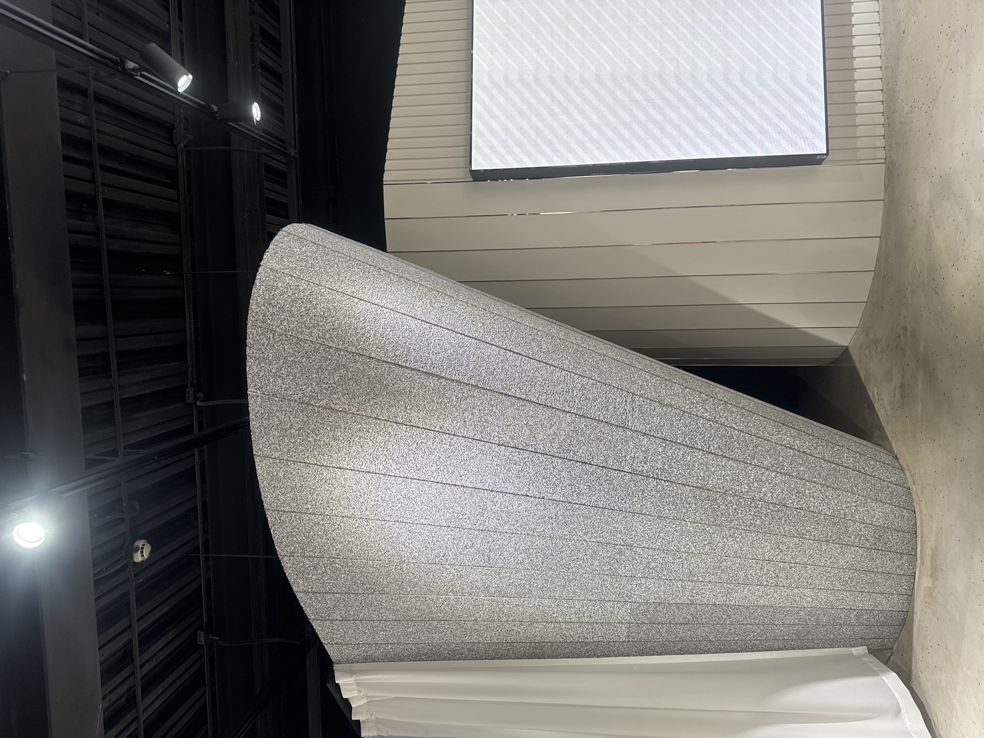 The image shows an interior space, possibly an exhibition or retail environment, with a distinctive curved, speckled gray structure. A white curtain hangs to the left of this structure. To the right, a vertical ribbed wall of a neutral color is visible, with a large, bright LED screen displaying a white patterned graphic. The ceiling above is black and industrial, with visible metal beams and track lighting. The floor appears to be concrete. The overall scene is modern and minimalist. This image was taken in Shanghai, China.