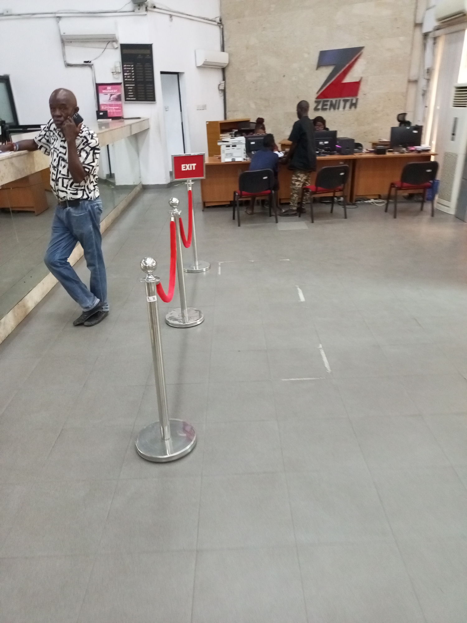 An interior shot of what appears to be a Zenith Bank branch in Satellite Town, Nigeria, showing a customer area and a service counter. In the left midground, a bald man wearing a patterned white and black shirt, blue jeans, and dark shoes, stands leaning against a counter while speaking on a black mobile phone. His reflection is visible in a reflective surface to his left.
In the center foreground, three chrome stanchions with red velvet ropes delineate a path, leading towards a red sign displaying "EXIT" in white letters. White tape markings on the light grey tiled floor indicate social distancing positions for customers.
In the background, a bank service counter is visible. A prominent "Z" logo and the "ZENITH" brand name are mounted on the wall behind the counter. Three individuals, presumably bank staff, are seated at workstations with computers and monitors, attending to customers. One customer, dressed in dark clothing and camouflage pants, stands facing a teller at the counter. Several empty black chairs with red backs are placed around the counter area. Air conditioning units and other administrative equipment are visible on the walls and near the counter. Part of a pink sign with the text "BE WARMED" is visible near the man on the phone. The scene is well-lit by indoor artificial lighting.