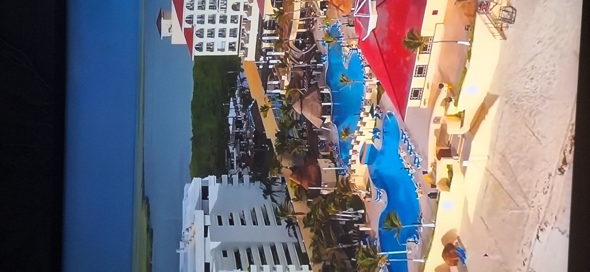 The image depicts an aerial view of a resort complex, possibly a hotel or condominium, situated near a body of water. Multiple buildings are visible, including a large white multi-story structure on the left and a building with a red roof on the right. The resort features several swimming pools, some with lounge chairs and umbrellas surrounding them. There are also thatched-roof structures, palm trees, and sandy areas that appear to be part of the beach. The environment suggests a tropical or coastal location. The sky is a deep blue, and there is a distant body of water and a strip of land with vegetation. No specific details about the city of Bila Tserkva, Ukraine are discernible in this image, as the scene appears to be a resort in a different geographical setting.
