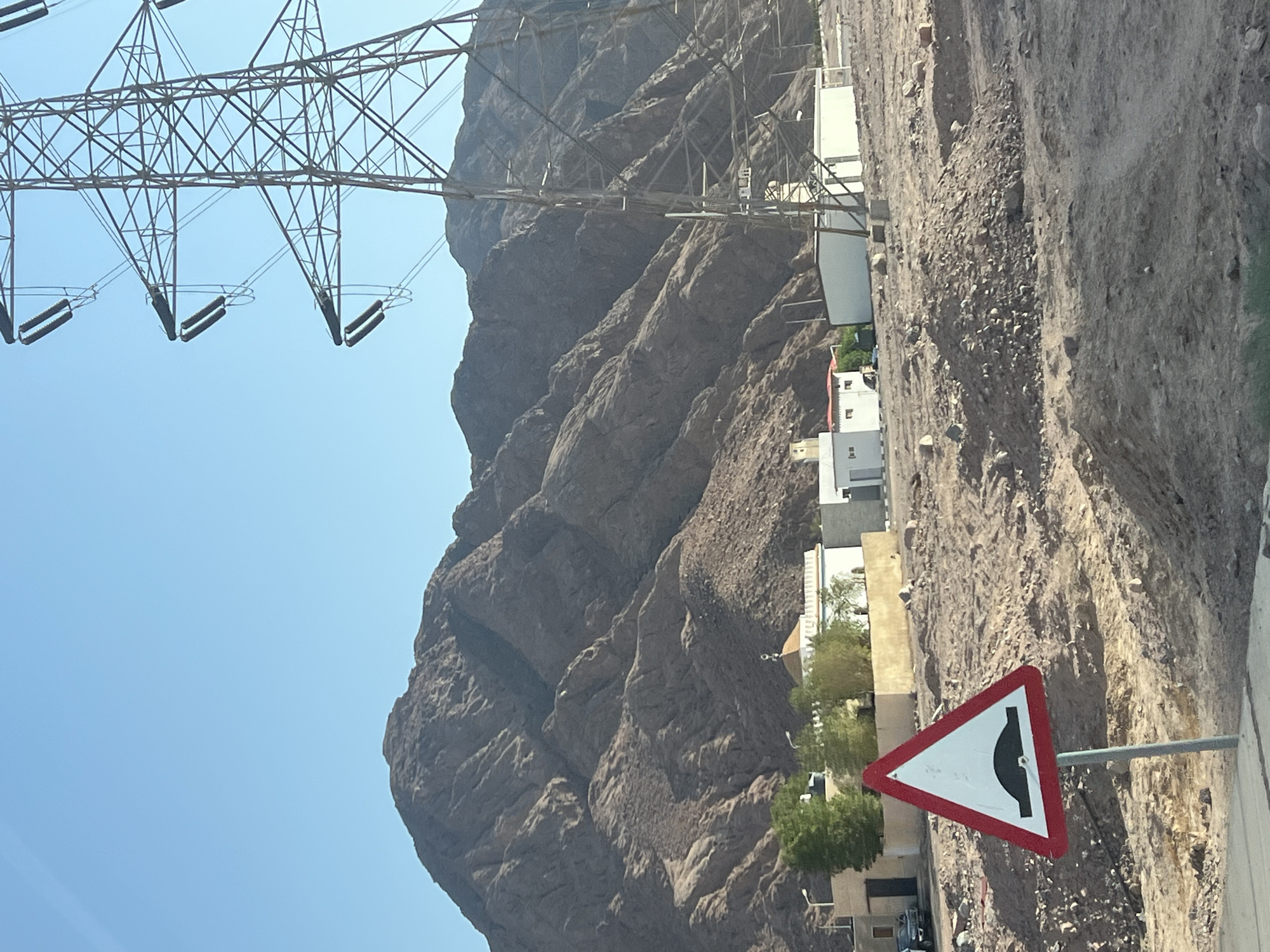 This image depicts a roadside scene in Dahab, Egypt, characterized by a stark, arid environment and human infrastructure. In the foreground, a red and white triangular speed bump warning sign is prominently displayed on a metal pole, indicating a potential hazard on the road. Behind the sign, a rocky, uneven terrain dominates the landscape, with scattered rocks and debris.
Further back, a cluster of modern buildings, appearing to be residential or small commercial structures, are visible. Some have white exteriors, and one features a domed roof, possibly a small mosque. Green trees and shrubs provide a touch of vegetation against the otherwise barren surroundings. A tall electricity pylon stands to the right of the frame, a testament to the region's power infrastructure. In the far background, imposing, rugged mountains rise under a clear blue sky, their slopes etched with the geological history of the area. The overall impression is one of a remote, developing settlement coexisting with the natural grandeur of the desert landscape.