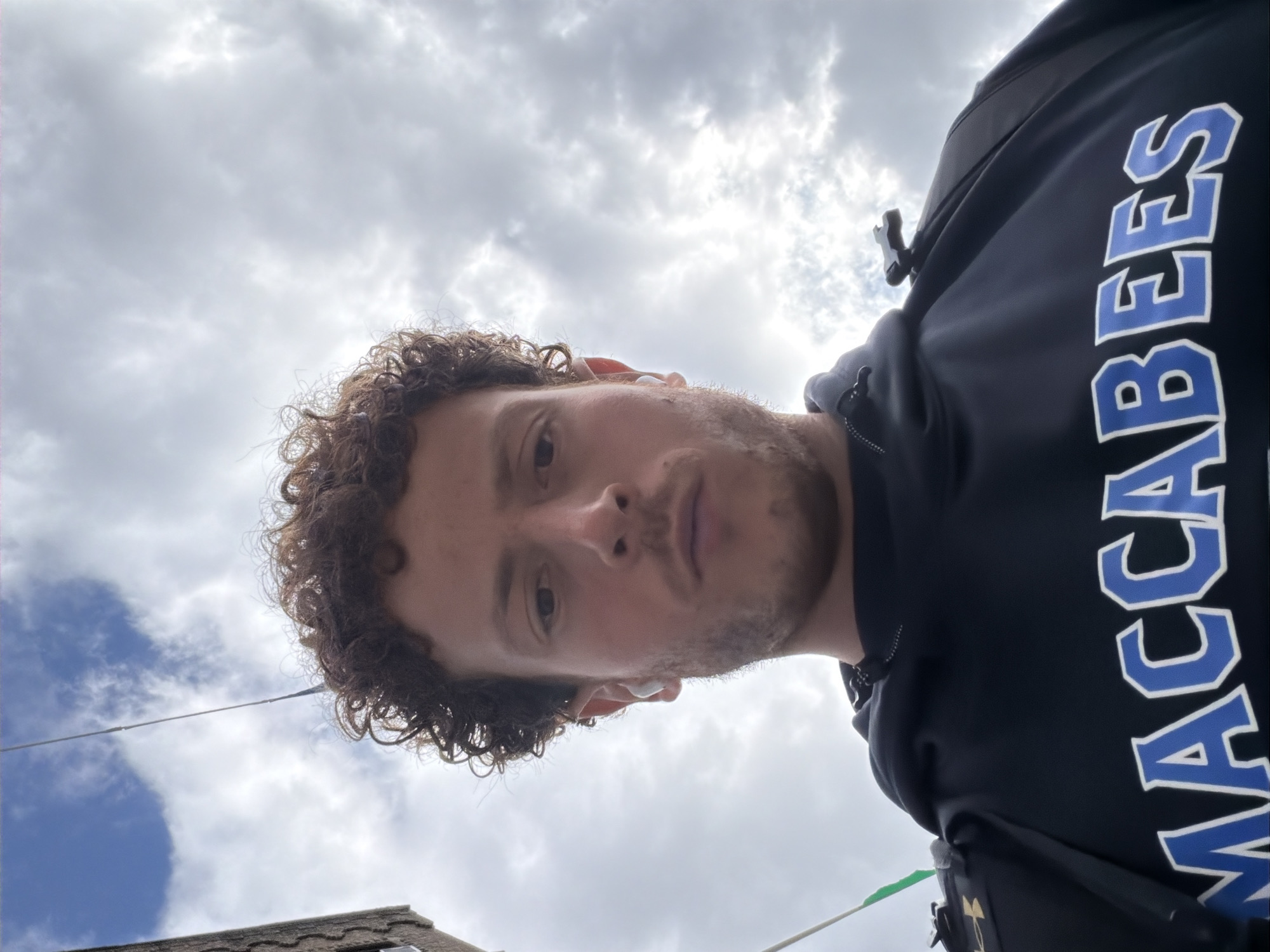 The image is a low-angle portrait of a young adult male, seen from the chest up, looking directly towards the camera. He has curly brown hair and a short beard or stubble. He wears a white earbud in his right ear. His expression is neutral.
He is dressed in a dark (likely black or navy blue) hooded sweatshirt. Prominently displayed on the right side of his chest/shoulder area is the word "MACCABEES" in large blue capital letters with a white outline. A small part of a strap, possibly from a bag, is visible near his left arm.
The background is dominated by a partially cloudy sky, showing patches of bright white clouds and some areas of blue sky, indicating daytime. In the lower left portion of the frame, a residential roofline is visible, suggesting an urban or suburban setting. A thin, dark wire or cable crosses horizontally through the lower half of the background. The overall scene suggests the person is outdoors, possibly in New York, United States of America.