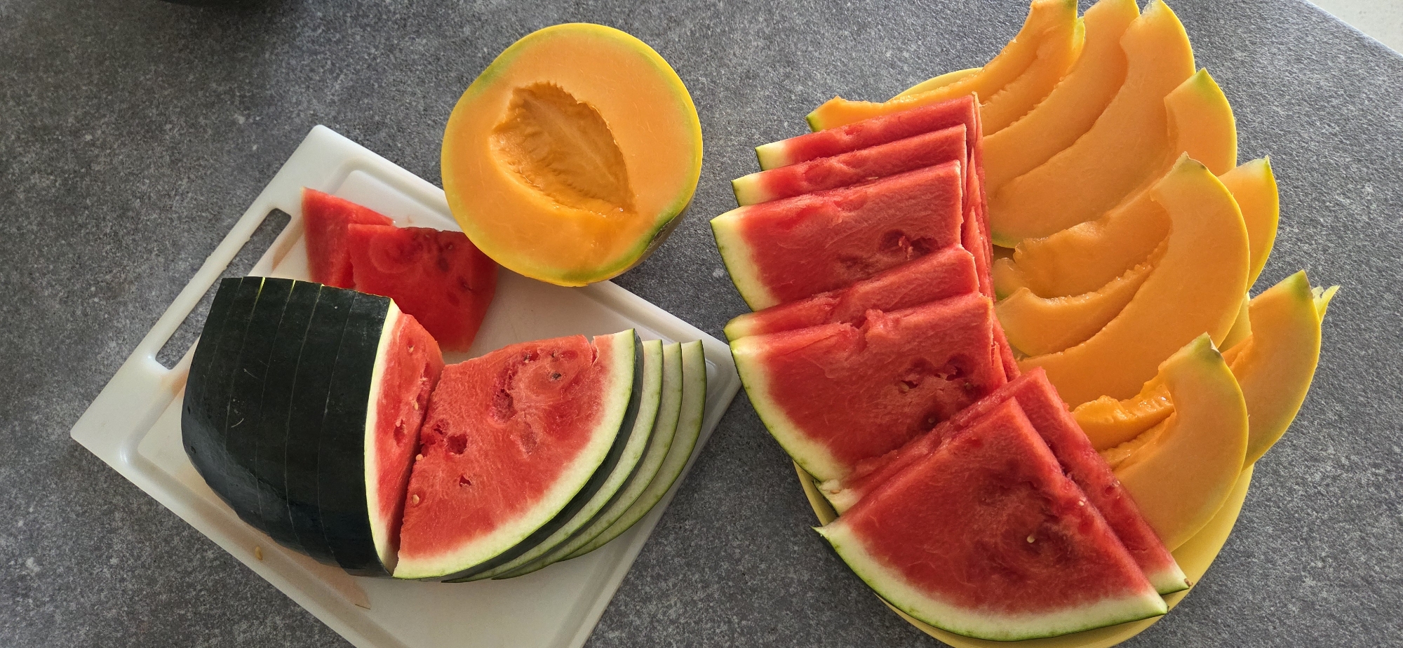 A high-angle, close-up shot shows a collection of sliced fruits, specifically watermelon and cantaloupe, arranged on a white cutting board and a yellow plate. The fruits are laid out on a speckled gray countertop. In the upper left, a partially sliced dark green watermelon is visible, with several fanned red slices and two smaller triangular pieces beside it on the cutting board. To the right of the cutting board, a halved cantaloupe with bright orange flesh and a scooped-out center rests on the countertop. Below, on the yellow plate, there are rows of fanned watermelon slices, overlapping with curved slices of cantaloupe. The scene is well-lit, highlighting the vibrant colors of the fruit. There are no people or animals present. The setting appears to be a kitchen or food preparation area. The location is not specified in the image.