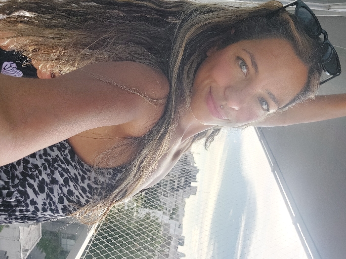 A fair-skinned woman with long, wavy brown hair smiles at the camera. She has a lip piercing and dark sunglasses rest atop her head. She is wearing a black and white patterned top. Behind her is a view of buildings, a body of water, and a sunny sky. The photo appears to be taken from a high vantage point, possibly a balcony.
