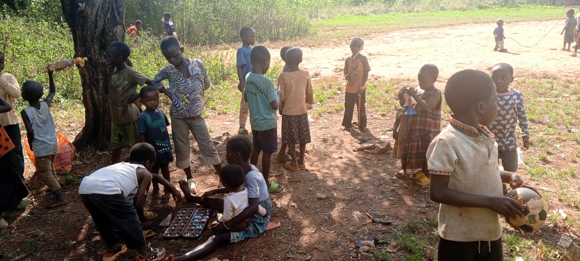 In Ngalula, Democratic Republic of the Congo, a group of children congregates outdoors on a dirt surface dappled with sunlight and shadows. Some children gather near a tree, one holding a doll. Several other children are positioned behind and to the right of the tree. In the center, a woman sits on the ground with a toddler in her lap, alongside a Mancala board game. A boy on the right holds a deflated soccer ball. Other children are scattered across the background. The surrounding environment includes trees, bushes, and a grassy area in the distance.