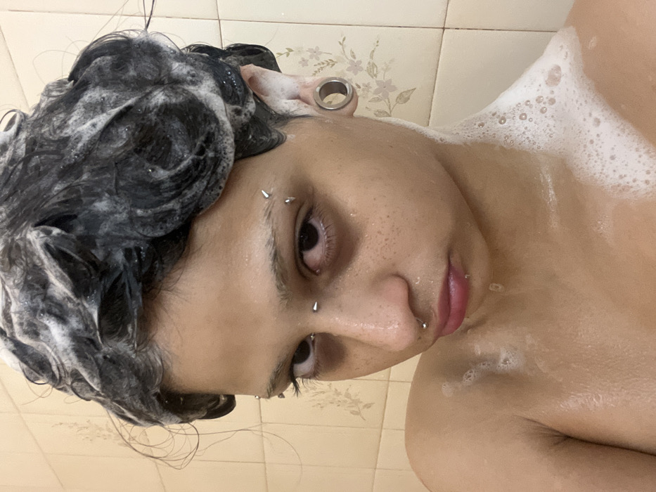 A close-up image shows a person in the shower, covered in soap suds. The focus is on their face, which has several piercings, including eyebrow, nostril, and lip piercings. Their dark hair is wet and coated with shampoo. An ear gauge is visible in the left ear. The background shows tiled shower walls. The person is looking slightly downward with a neutral expression.