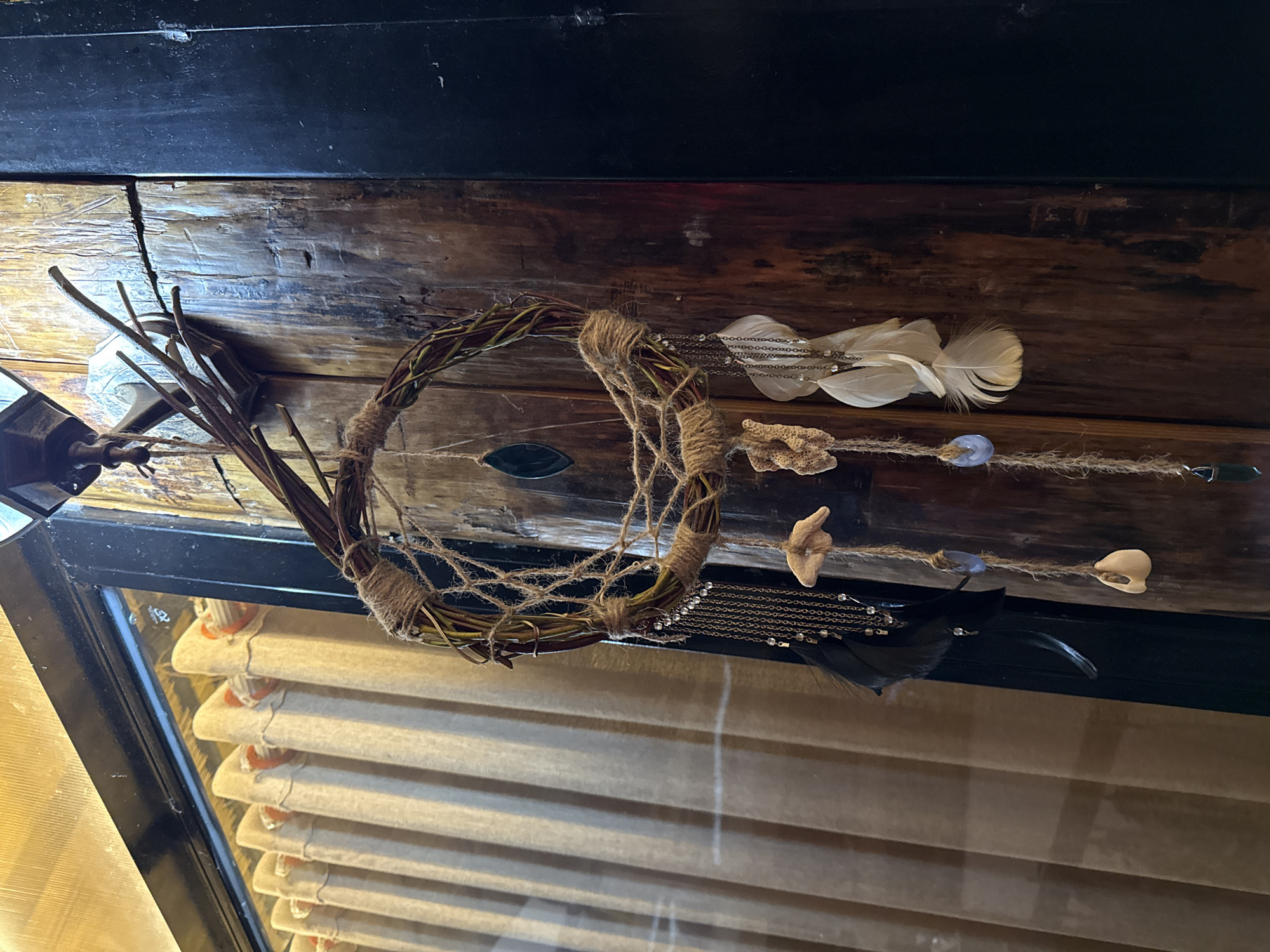 A dreamcatcher made of twigs and twine hangs against a brown wooden surface. It features white and black feathers, beads, a shell, and a piece of coral. The dreamcatcher is in front of a window with closed blinds. The location is presumably in Bac Natural Heritage Area Trang, Vietnam.