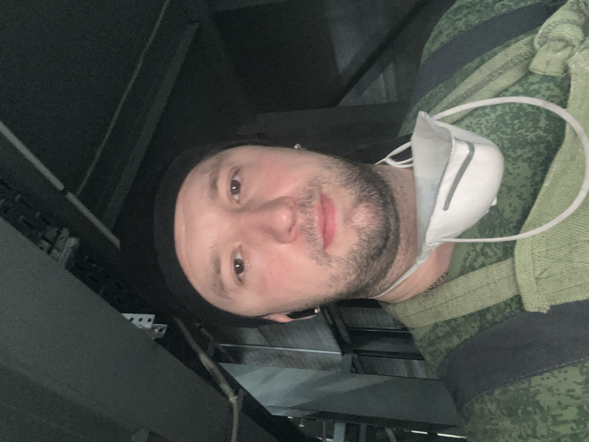 A man wearing a camouflage jacket and a black beanie is looking into the camera. A white face mask hangs from his neck. The background is dark and appears to be an industrial setting with metal structures and cables. The location is Noviy, Russia.