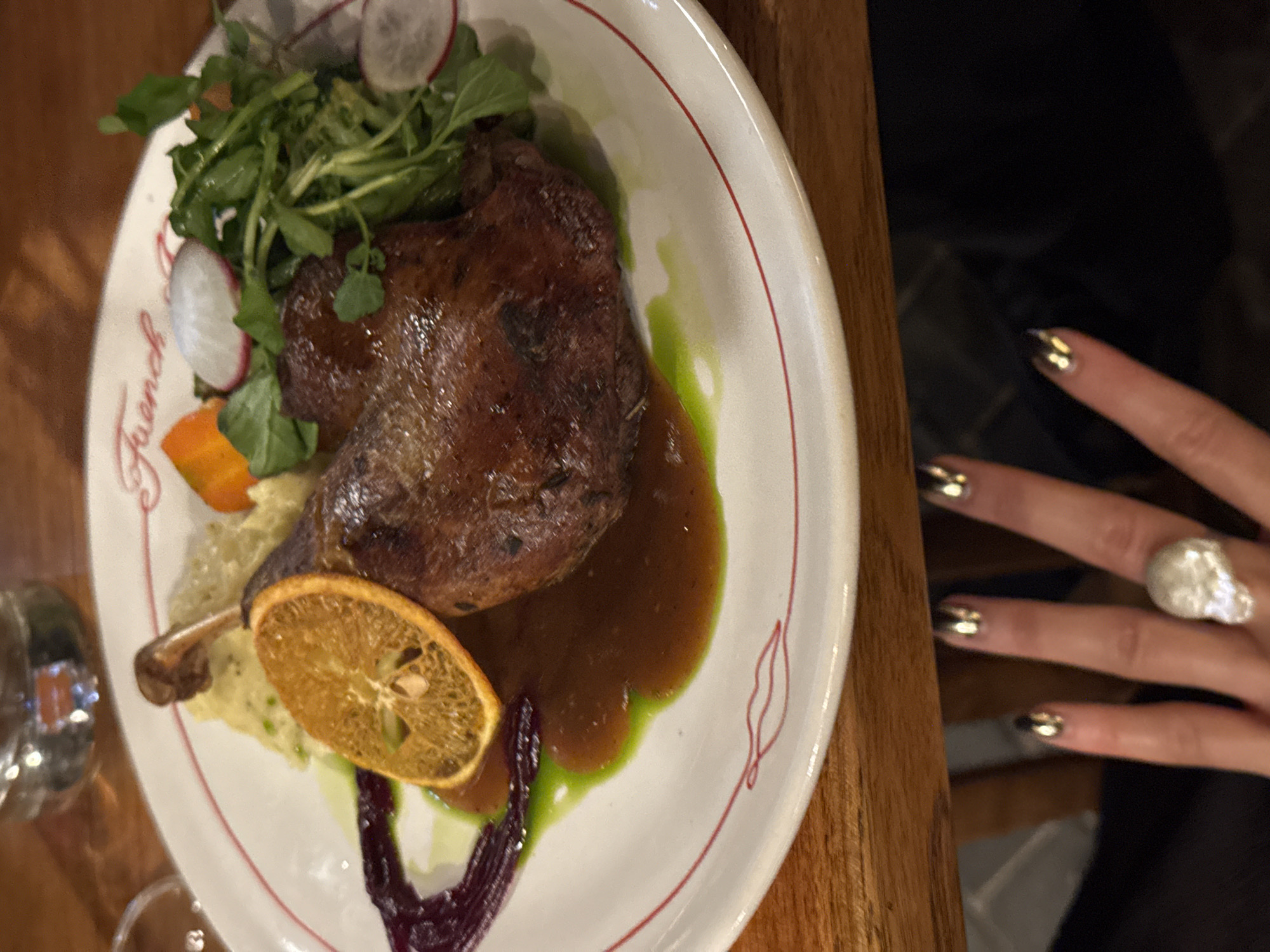 In the city of Ubud District, Indonesia, a close-up shot captures a plate of duck confit. The dish is presented on a white plate with a red rim, featuring a portion of mashed potatoes, a slice of dried orange, a vibrant green sauce, and a garnish of arugula and thinly sliced radishes. The duck itself is a deep brown, glistening with sauce. The plate is positioned on a wooden table, with a blurred background suggesting an indoor dining setting. A person's hand, adorned with metallic nail polish and a prominent silver ring shaped like a skull, is visible in the foreground, reaching towards the plate. The overall impression is one of a meticulously prepared and artfully presented meal.