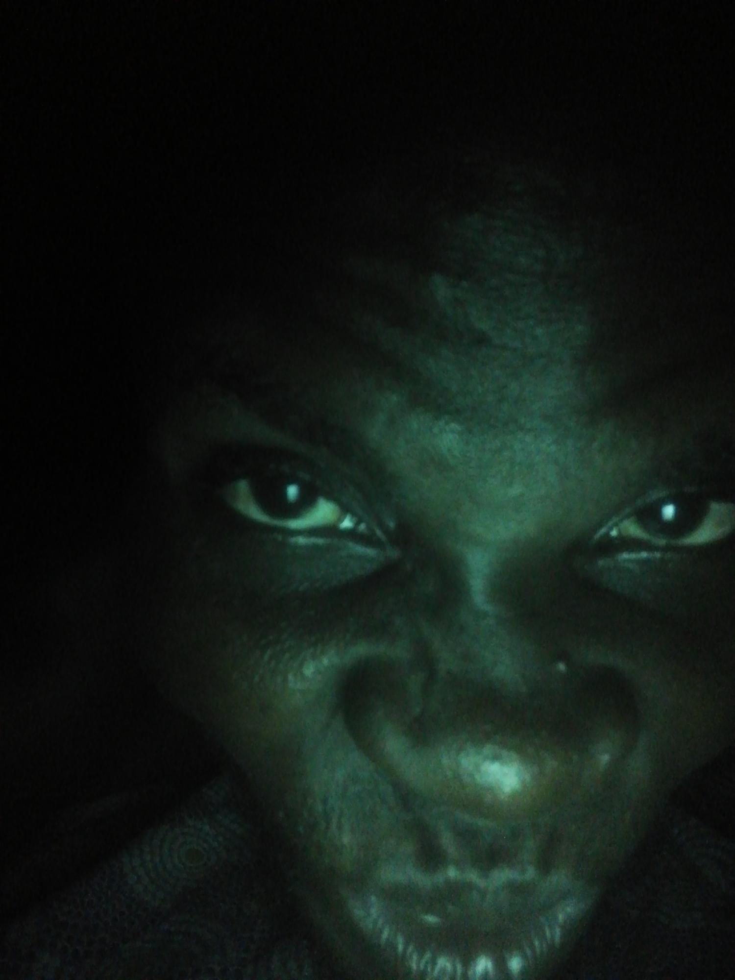 A close-up, low-light image shows a person's face. The person appears to have dark skin. The eyes are open and facing forward. The nose is prominent, and the mouth is slightly open. The environment is dark and unclear.