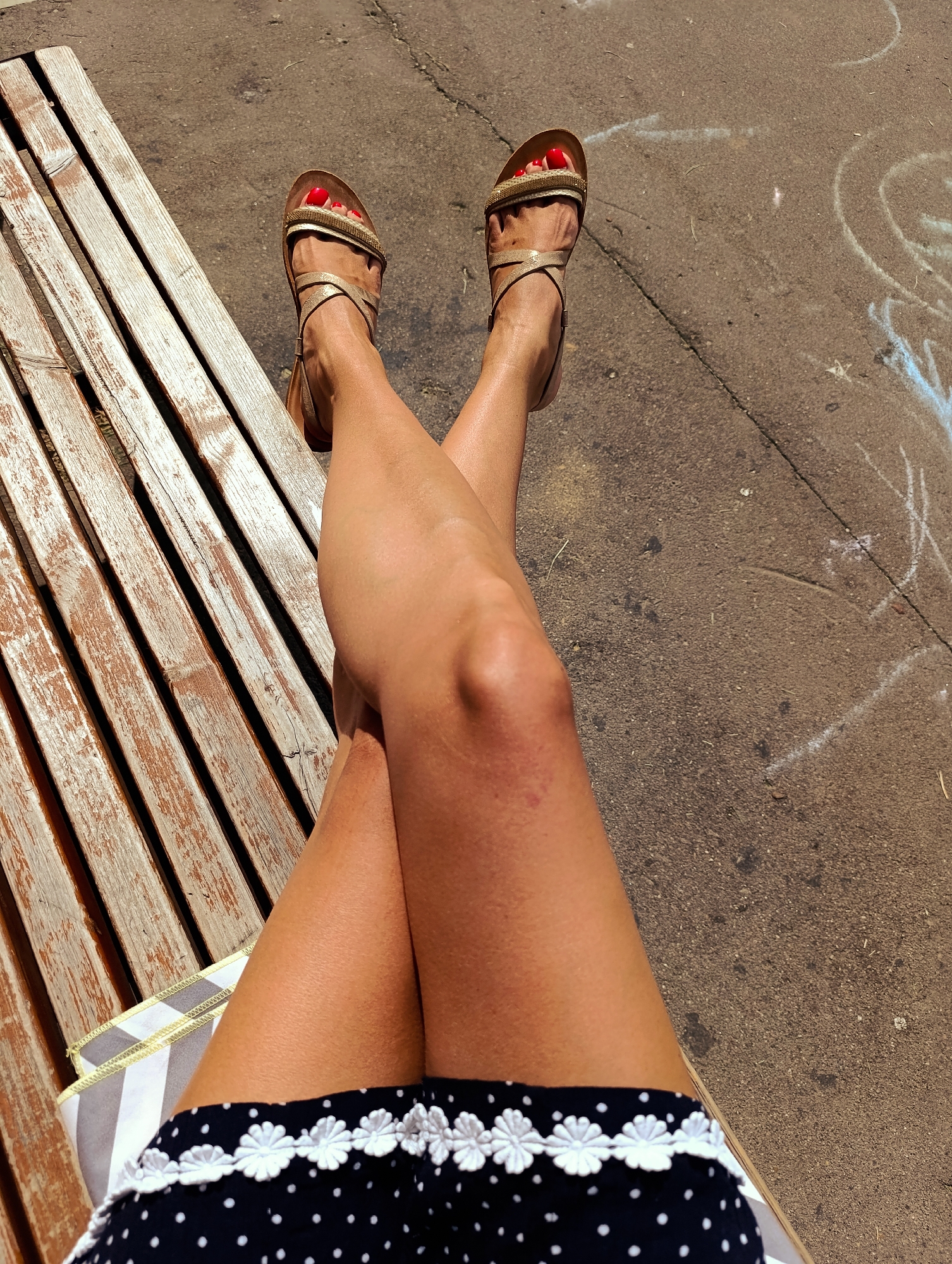 A first-person perspective image shows a person's legs and feet, adorned with red nail polish and gold sandals, crossed while sitting on a weathered wooden bench. The bench is positioned on a paved surface, likely a sidewalk or park pathway, with chalk markings visible in the background. The scene appears to be outdoors during the day, with natural lighting casting shadows. The overall impression is one of casual relaxation in an urban outdoor setting. It is not possible to determine the specific location in Moscow, Russia from the provided image.