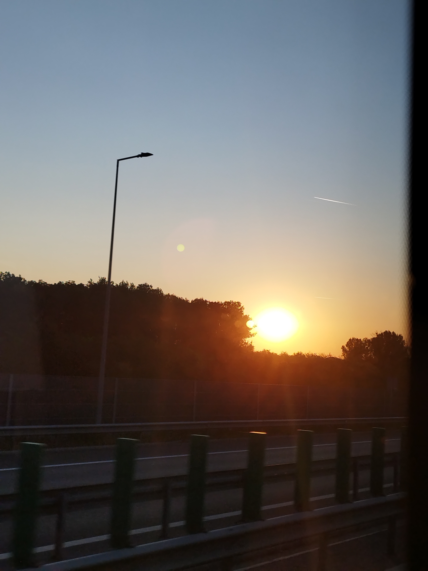 The image captures a sunset over a highway in the vicinity of 1 Decembrie, Romania. The sun, a bright orange orb, is partially obscured by a dense line of trees that form a dark silhouette against the sky. The sky transitions from a pale blue at the top to a warm orange and yellow hue near the horizon. A single streetlamp, silhouetted in black, stands tall to the left of the frame. In the sky, two faint white contrails from an airplane are visible. Below the tree line, a highway is visible with a guardrail. The highway appears to be empty of vehicles. Green posts line the edge of the roadway. The overall lighting suggests the golden hour of sunset. The scene is viewed from a slightly elevated perspective, possibly from within a moving vehicle.