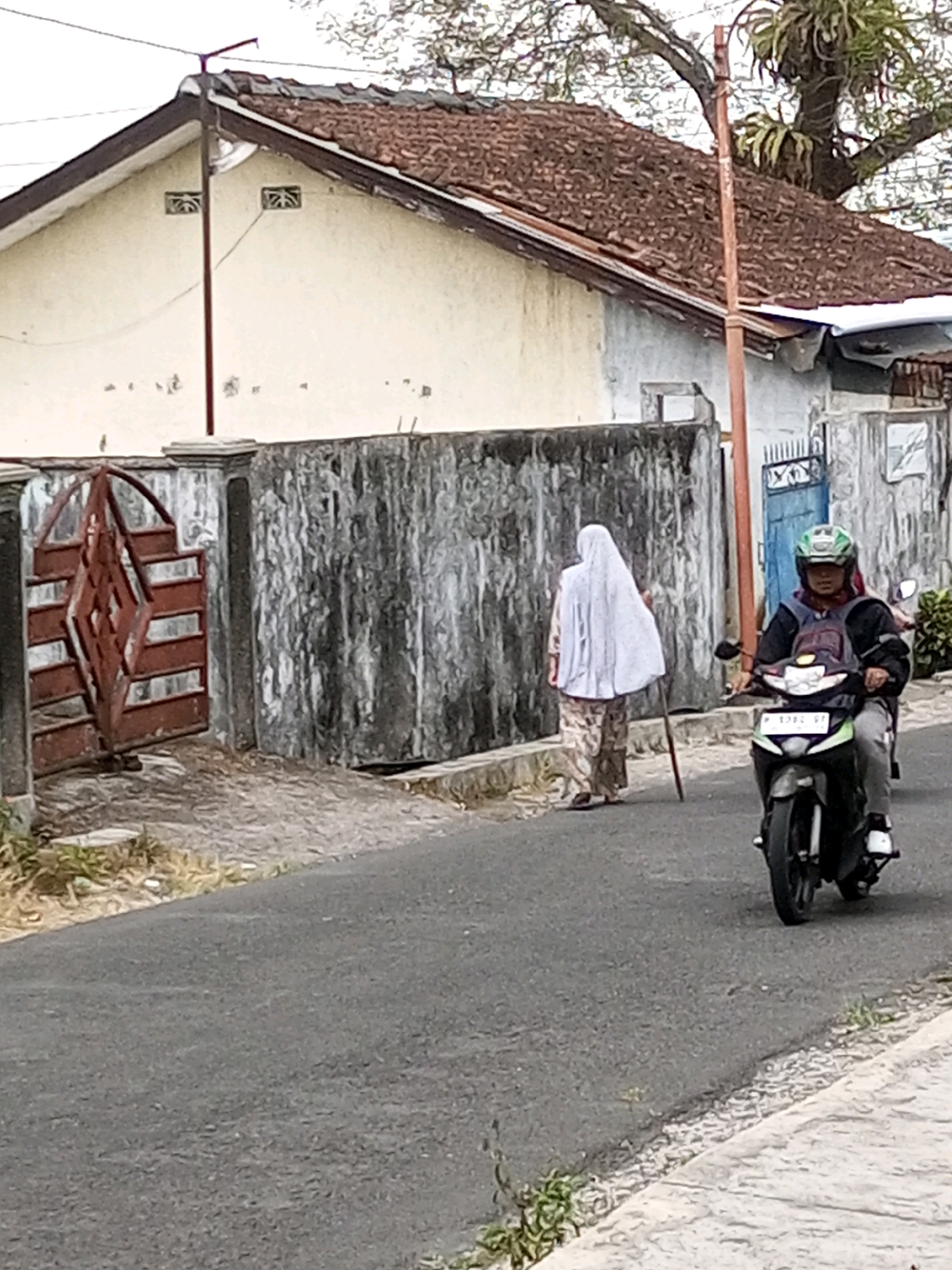 The image captures a street scene in Krasak, Indonesia. In the foreground, a person wearing a green helmet and dark jacket rides a motorcycle. The motorcycle has a license plate with illegible characters. Behind the motorcycle, an elderly person walks away from the camera, draped in a white headscarf and patterned clothing, using a walking stick for support. They are walking along a road adjacent to a weathered, textured wall. To the left of the wall is a red metal gate with a diamond pattern, partially open. The background features a building with a terracotta tiled roof and light-colored walls, with some vegetation visible. The overall atmosphere is of a typical day in a residential area.
