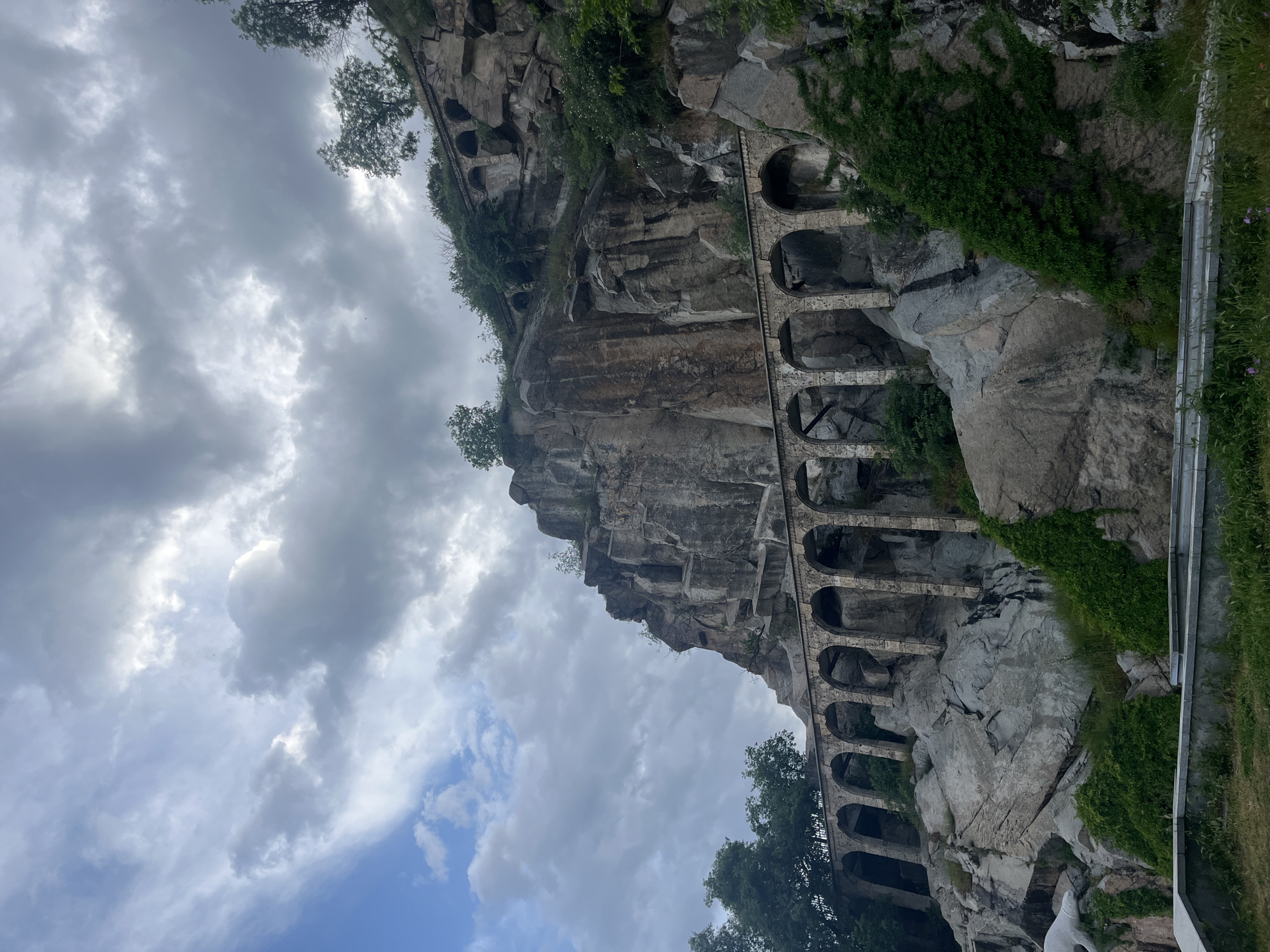 The image depicts a rock face with a series of arched structures built into it. These structures appear to be part of a road or walkway carved into the rock. Vegetation, including trees and vines, grows on and around the rock face and the arched structures. Above the rock face, there is a cloudy sky. A guardrail is visible on the right side of the image, suggesting this location is near a road.
Based on the visual evidence of architectural style and vegetation, this image may depict part of the "Roman road" or "Roman Aqueduct" within the "Three Hills" area in Plovdiv, Bulgaria. The image seems to show a section that resembles a Roman aqueduct, although the visible structure may have served another purpose. The architectural style of the arches and the way they are embedded within the rock face is consistent with that of Roman-era construction.