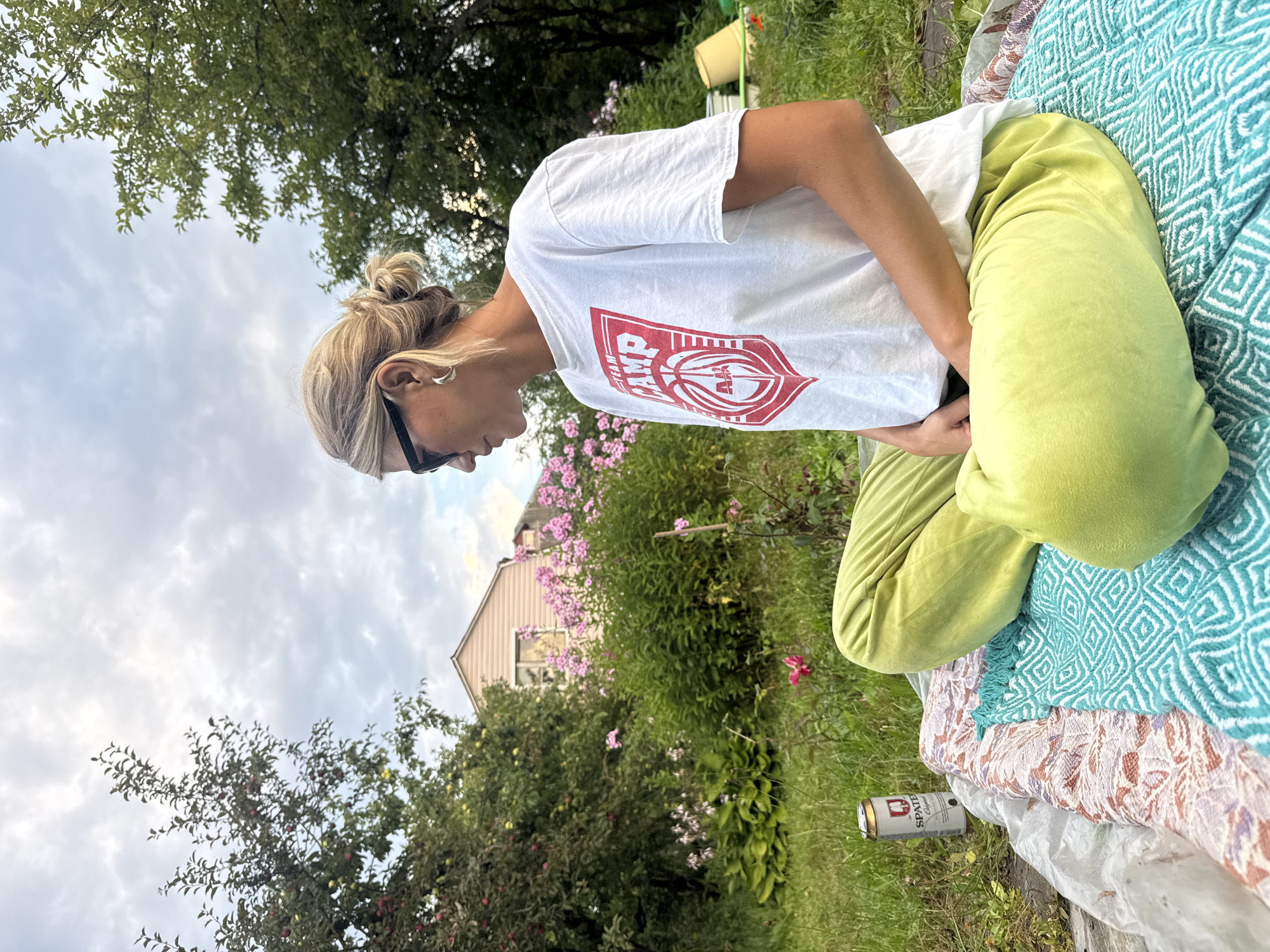 In the city of Krasnaya Presnya Snt, Russia, a woman with blonde hair pulled back in a bun is sitting cross-legged on a blanket in a garden. She is wearing a white t-shirt with a red graphic and loose-fitting light green pants. She has on black sunglasses and silver hoop earrings. In the background, there are trees, pink flowers, and a light-colored house. A can of "SPATE" beer is on the blanket next to her. The sky is overcast.