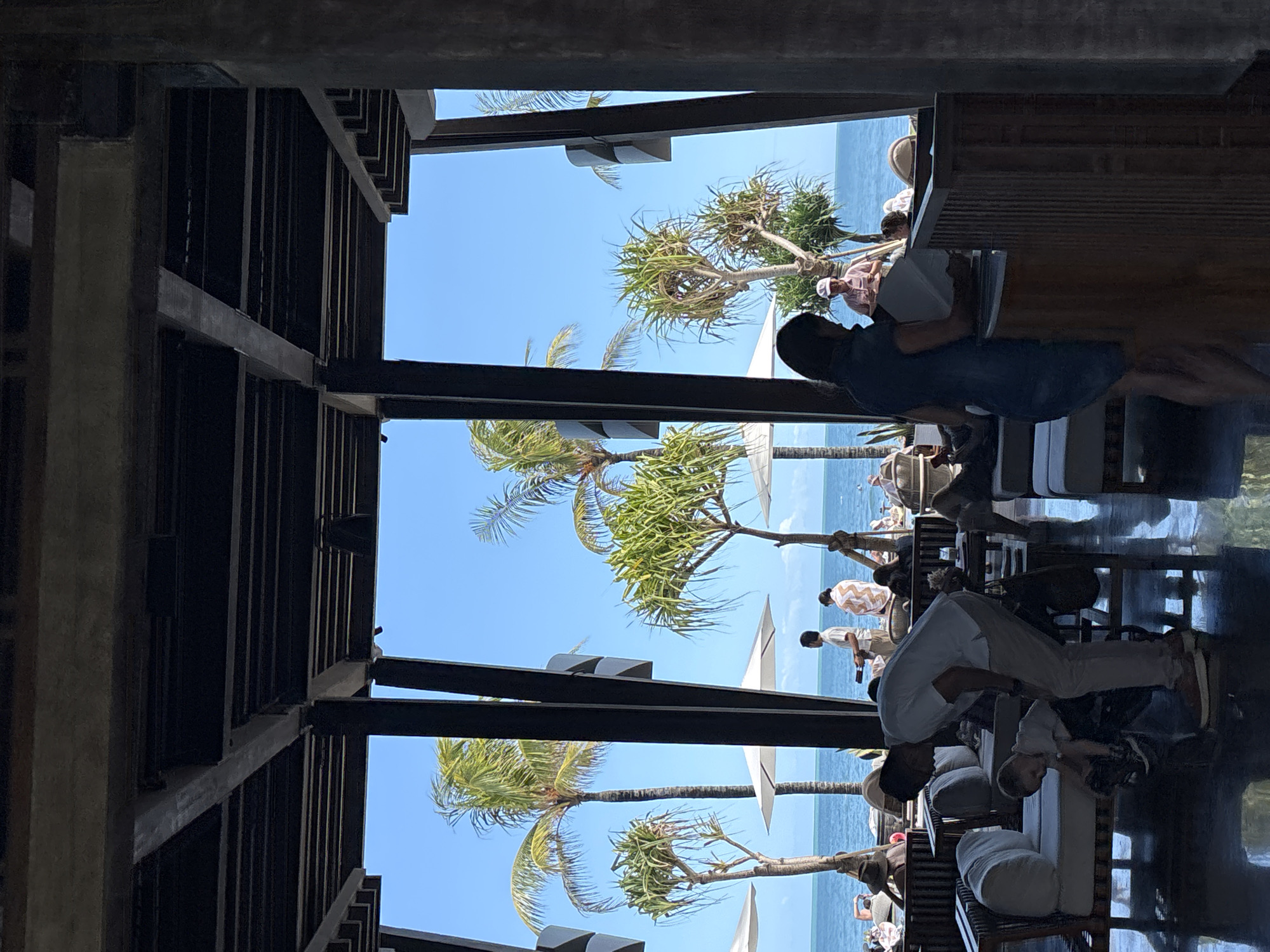 The image shows a partially obscured view from inside a structure, likely a resort or open-air building, in North Kuta, Indonesia. The view is oriented towards the ocean.
**People:**
* Several individuals are present in the outdoor area. Some appear to be relaxing on lounge chairs, likely guests or patrons.
* A person is sitting at a table, possibly reading or working.
* Two adults are together, possibly a parent and child, with the child interacting with something on a table.
**Objects:**
* Lounge chairs and tables are positioned outside.
* The structure features a dark wooden or metal framework, providing shade and defining the space.
**Environment:**
* The scene takes place in North Kuta, Indonesia, evidenced by the tropical vegetation (palm trees) and ocean view.
* The sky is clear and blue.
* The overall setting is a luxurious or resort-like environment.