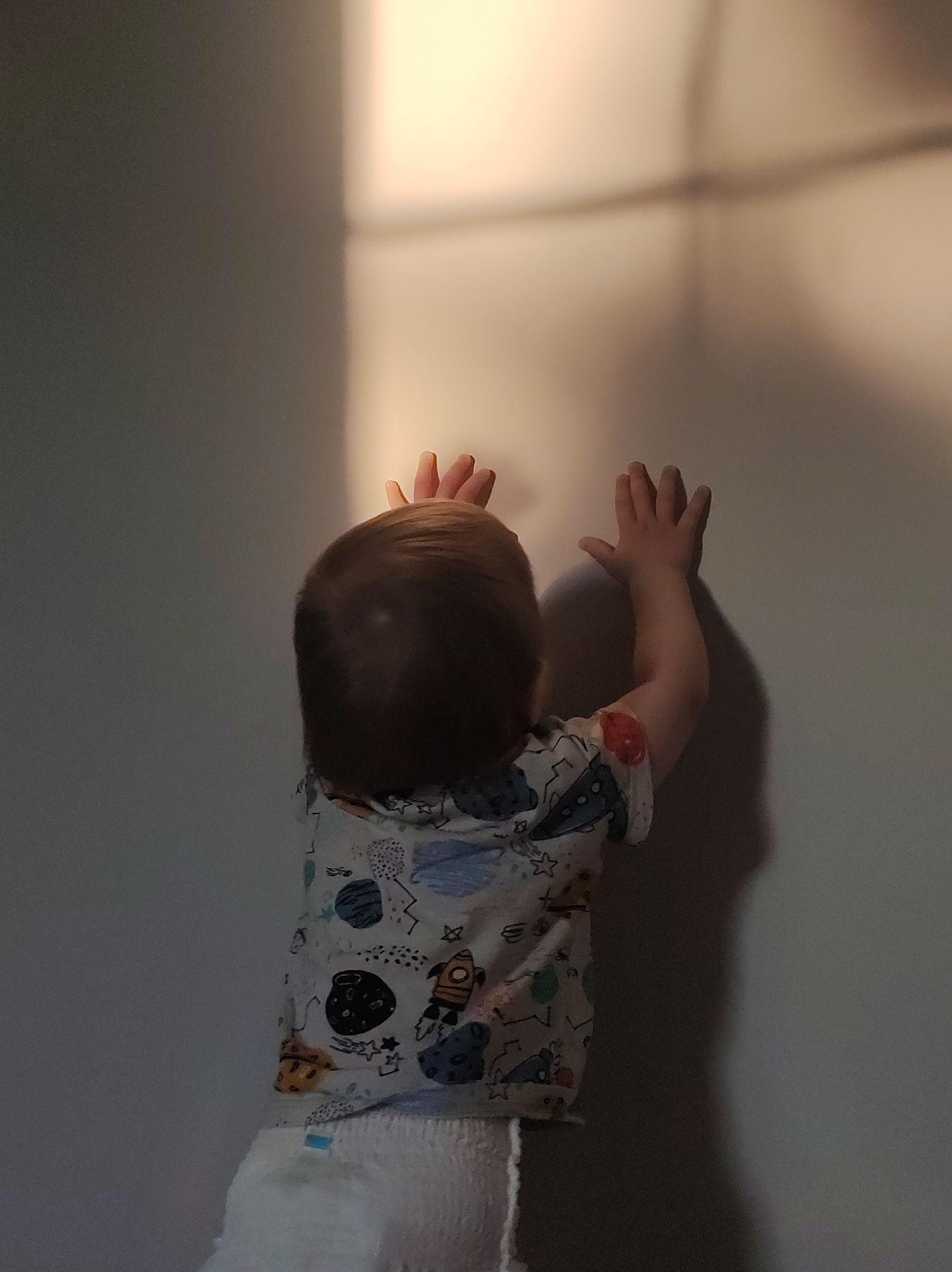 A child stands with their back to the camera, reaching up towards a wall. The wall is illuminated by a square patch of light. The child is wearing a printed shirt and a diaper. The location appears to be indoors. The city and specific location are unknown.