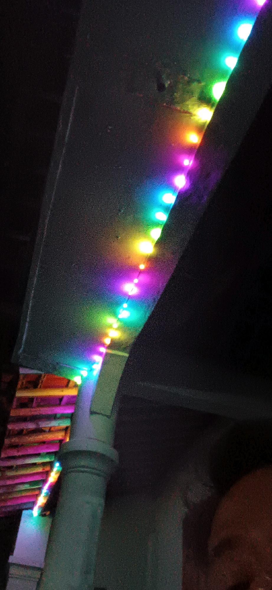 In Wonokerso, Indonesia, an image captures a string of colorful LED lights illuminating a dark, outdoor environment. The lights, displaying a spectrum of colors from purple to green and yellow, are draped along an angled surface. Below this, a cylindrical, light-colored object, possibly a pillar or pipe, is visible. Further down, a row of wooden planks, also illuminated by the lights, can be seen. The background is predominantly dark, suggesting nighttime or a dimly lit area. A portion of a person's head and shoulder, with dark hair and skin, is visible in the lower right corner, partially obscured. The overall scene appears to be taken from a low angle, looking upwards.