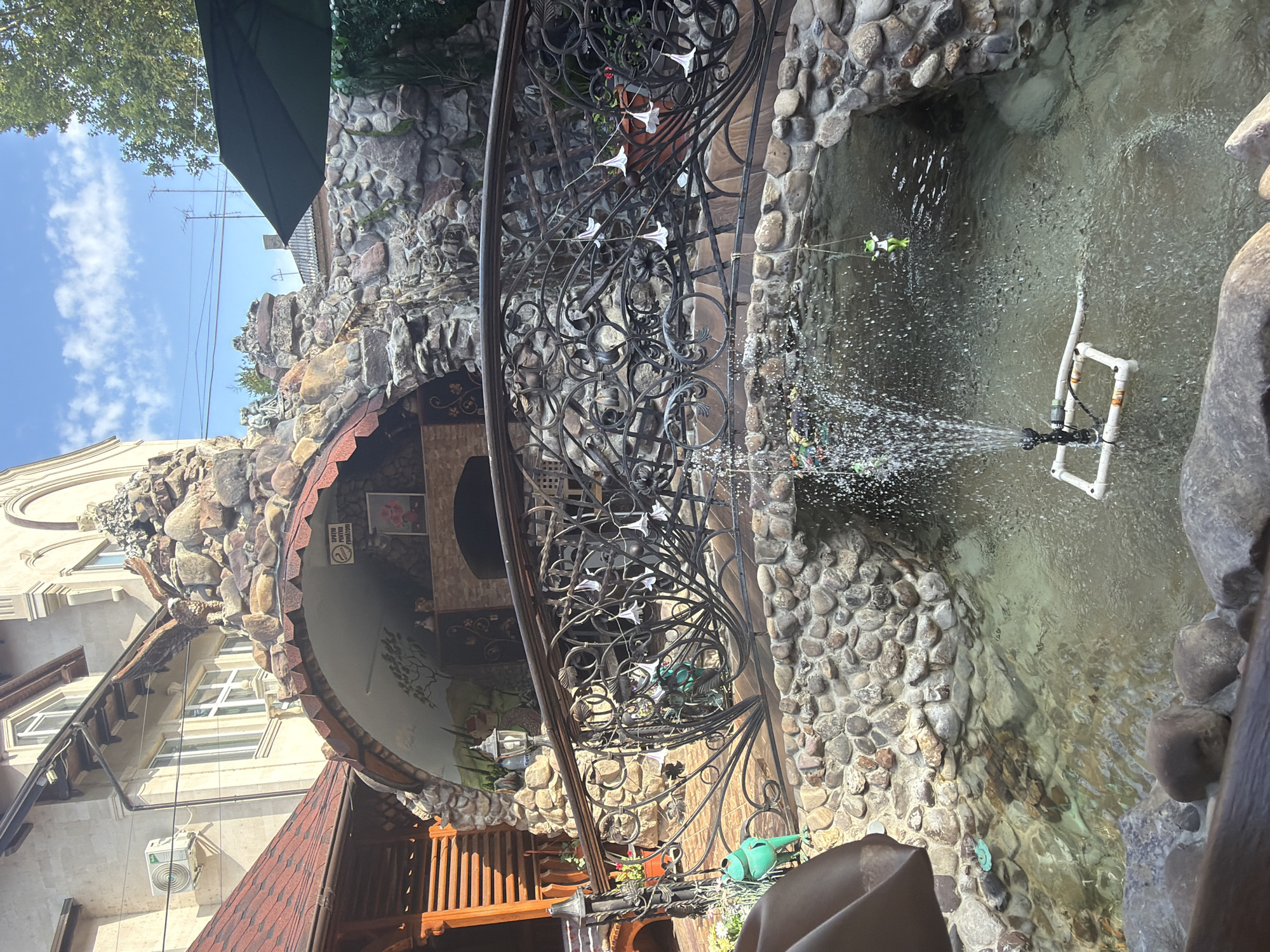 The image shows an outdoor fountain area with a bridge and a building. The fountain has a spray of water in the center, with a small white pipe system at its base. The water is clear and shallow, with rocks visible beneath the surface. The bridge is made of dark metal with intricate scrollwork and several small white flowers hanging from it. It spans over the fountain, connecting two stone-covered structures. To the left of the fountain, a green ceramic frog statue is positioned on the edge of the bridge. Behind the bridge, a building with a beige facade and a dark red tiled roof is visible. Part of the building features an arched entryway decorated with rocks and a mural. Above the arch, a large rock formation is present, topped with what appears to be a statue of an eagle. An air conditioning unit is mounted on the left side of the building's facade. A green umbrella is partially visible in the upper right corner of the image. The sky is blue with scattered clouds. The overall scene appears to be a garden or courtyard area, possibly part of a residential or hospitality establishment. The text on a sign above the arched entryway translates to "Smoking Area". The image is likely taken in Soroca, Moldova, given the mention of the location. There are no people visible in the image.