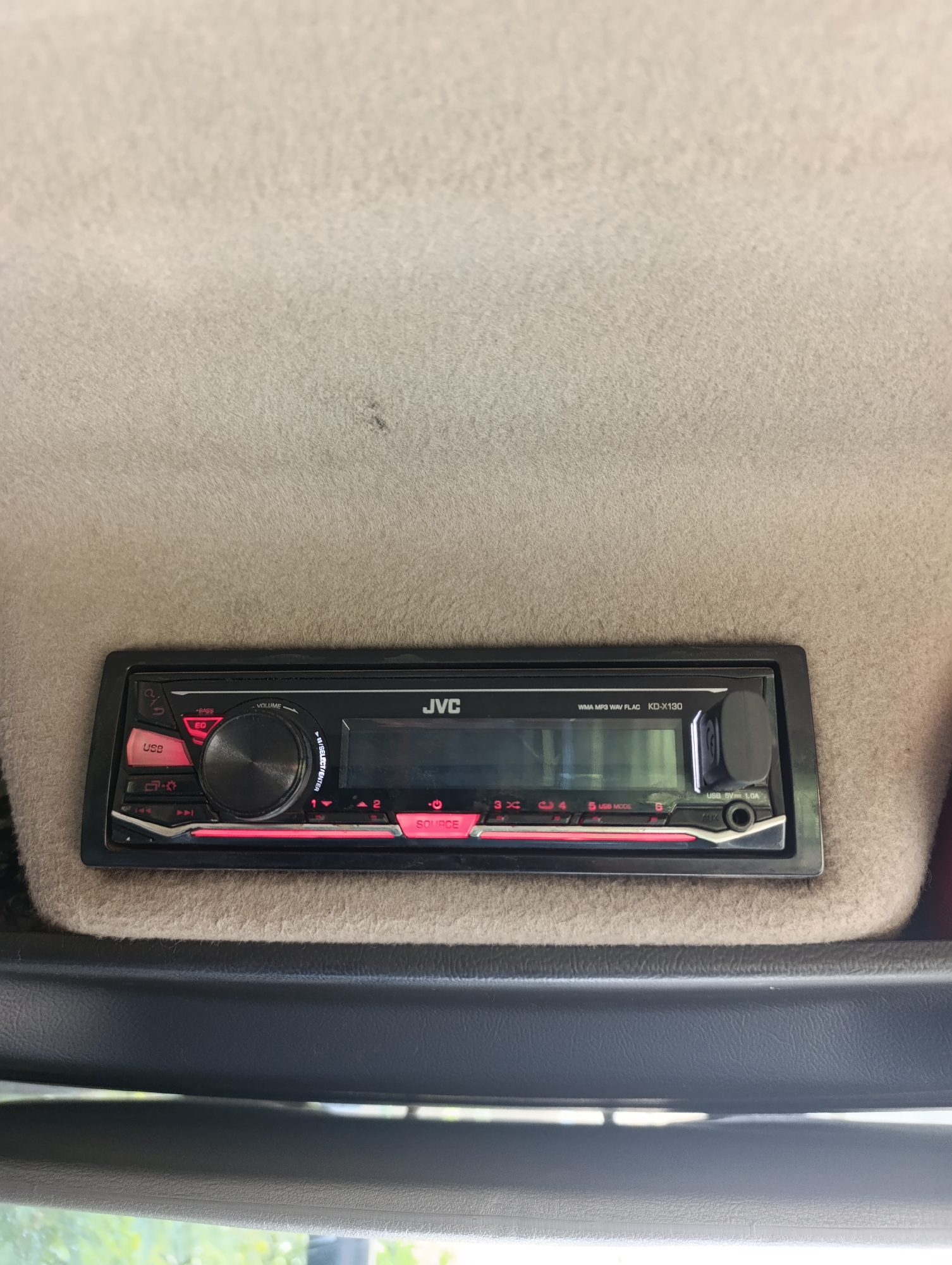An in-car JVC KD-X130 stereo system is mounted in a light beige upholstered dashboard. The stereo has a black faceplate with a digital display. Buttons are illuminated in red and include "EQ", "USB", and "SOURCE". A prominent black rotary knob is present for volume control and selection. A USB port and an auxiliary input are visible on the lower right of the faceplate. The stereo is positioned above a darker grey textured plastic trim. The background is entirely the textured, light beige upholstery of the car's interior. The image was taken in Dnipro, Ukraine.