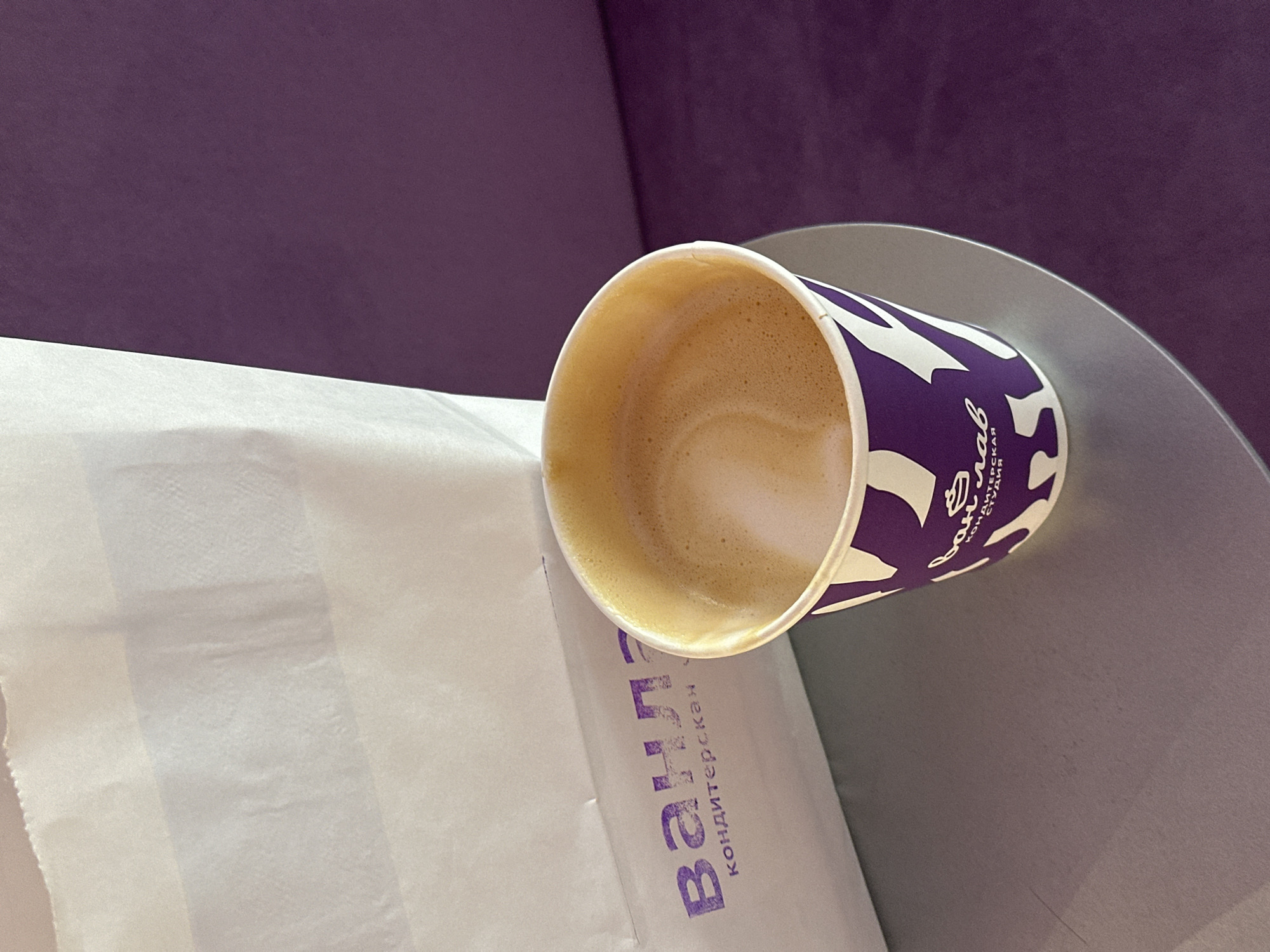 A coffee cup from "Vanyla," a confectionery in Kazan, Russia, sits on a grey table next to a white paper bag. The cup is mostly full, revealing a tan-colored coffee with a foamy top. The exterior of the cup is white with purple graphics. "Vanyla" is printed in Cyrillic on the bag. The background is a purple, upholstered surface.