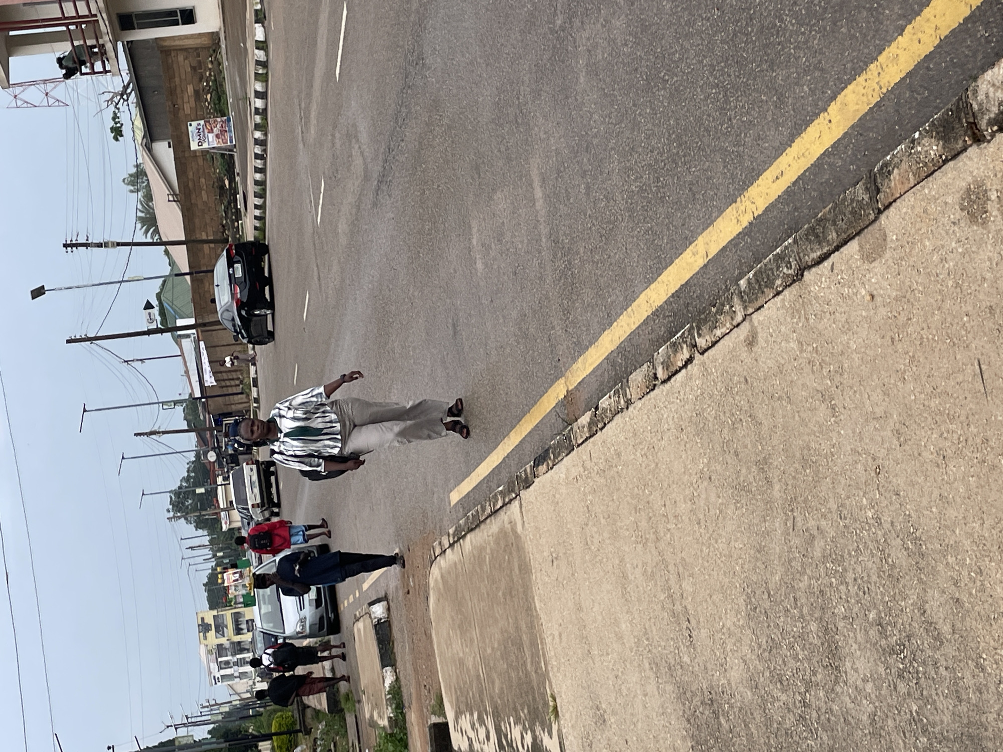 In the city of Jos, Nigeria, on a cloudy day, several people are seen walking along the side of a paved road. A woman in the foreground, wearing a striped shirt and light-colored pants, is walking towards the right side of the frame. Behind her, a black sedan is driving on the road. Further down the road, a group of young men are walking in the same direction as the woman. One of them, wearing a red shirt and blue shorts, has a backpack. To the left of the road, a sidewalk is visible, with some patches of greenery. In the background, buildings and streetlights can be seen, indicating an urban environment. The road has a yellow line marking the lane division.