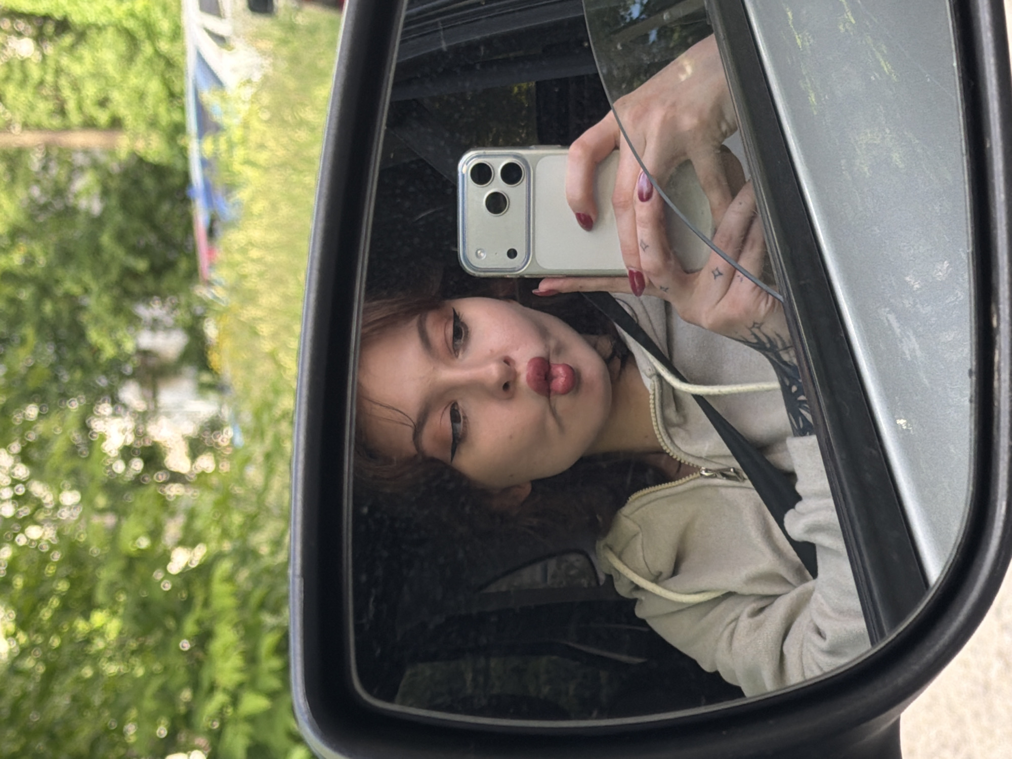 A young woman with dark curly hair and bold eyeliner is taking a selfie in a car's side mirror. She is wearing a light beige hooded sweatshirt and has her lips puckered. Her left hand, adorned with dark red nail polish, holds a silver smartphone with a three-camera setup. A tattoo of stars is visible on her right forearm. The background reflected in the mirror shows lush green foliage, suggesting an outdoor setting, possibly in a residential area or along a tree-lined street. The car's interior, including a seatbelt, is also visible. The overall scene is casual and playful.