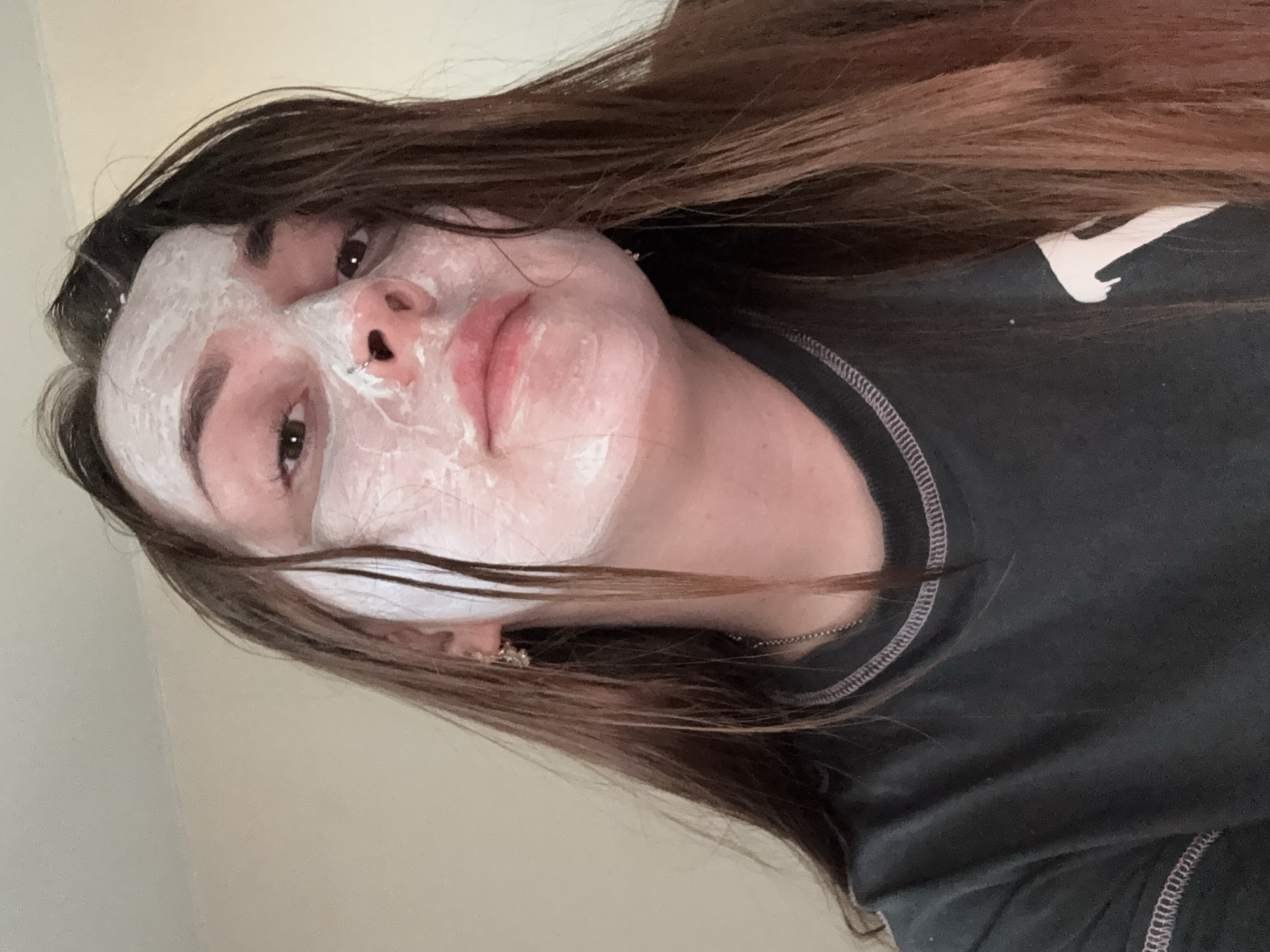A young woman with long brown hair has a white facial mask applied to her face. She is wearing a dark-colored shirt. The shirt has a white logo on it. The image is taken indoors. There is no information about Izhevsk, Russia.