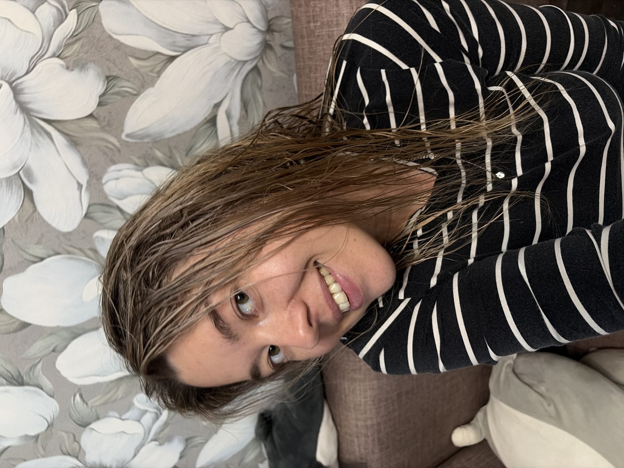 An indoor, upward-angled shot captures a smiling young woman, likely in her 20s or 30s, in what appears to be a home in Chaikovsky, Russia. Her light brown hair is visibly wet, with individual strands clinging together. She is wearing a black and white striped long-sleeved top.
The woman's head is tilted, and she looks directly towards the camera with light brown eyes, displaying a wide, cheerful grin that reveals her white teeth. Her expression conveys a sense of happiness and relaxation.
The background features a wallpaper with a repeating pattern of large, stylized white flowers, possibly magnolias, intertwined with grey-green leaves against a light grey or beige backdrop. To her right and partially obscured, a brown upholstered surface, likely a sofa or armchair, is visible. A soft, light-colored object, possibly a pillow or stuffed animal, can also be seen near her arm. The overall setting suggests a casual and comfortable home environment. There is no visible text in the image.