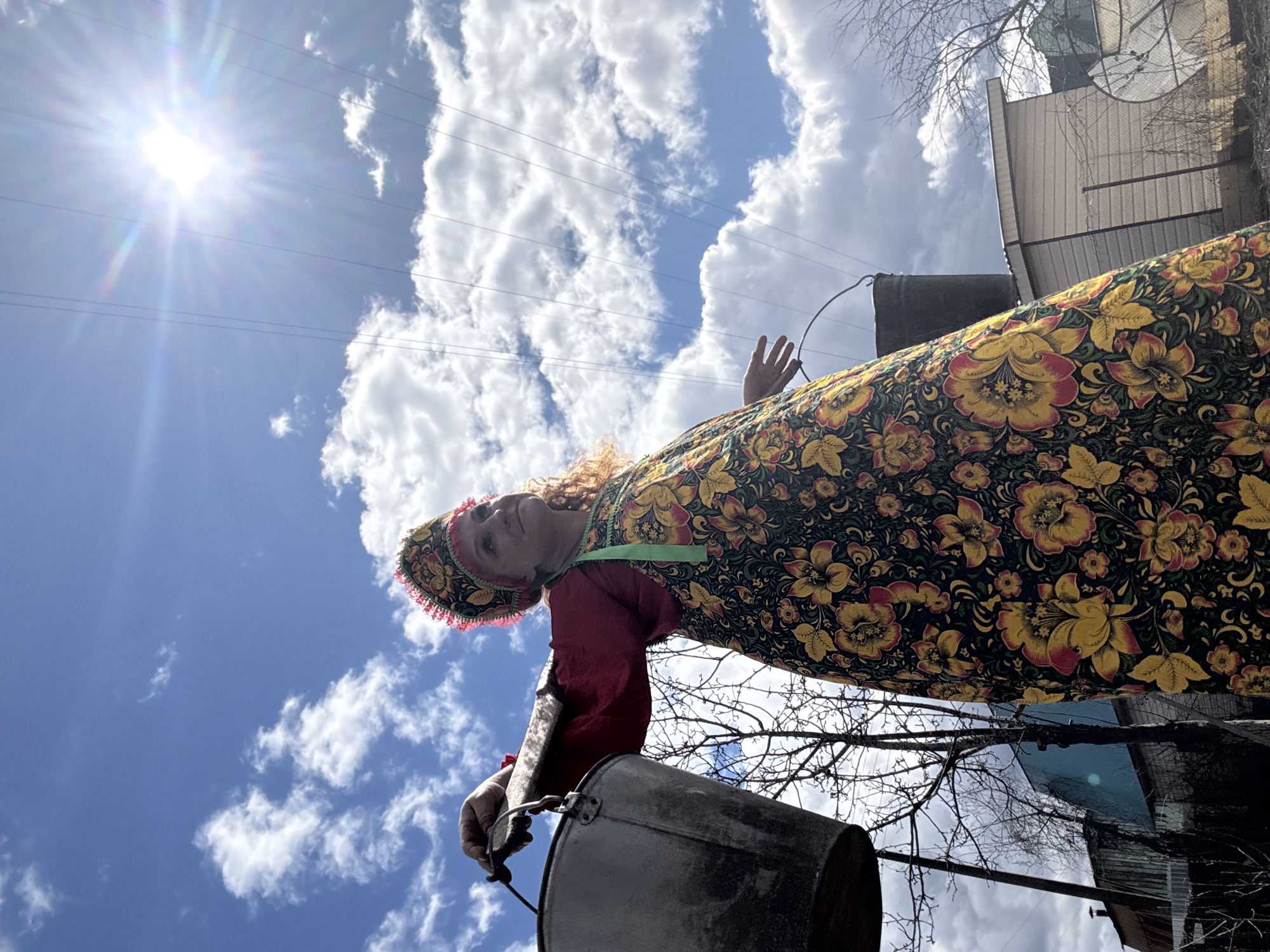 A low-angle shot depicts a person holding a metal bucket against a backdrop of a blue sky with scattered clouds and a bright sun. The individual is wearing a traditional Russian floral patterned dress and head covering, with a red underdress and green ribbon. The person's left hand is raised toward the sky. Bare tree branches are visible around the edges of the frame. In the background, there is a building with a metallic roof. This image was taken in the city of Chaikovsky, Russia.