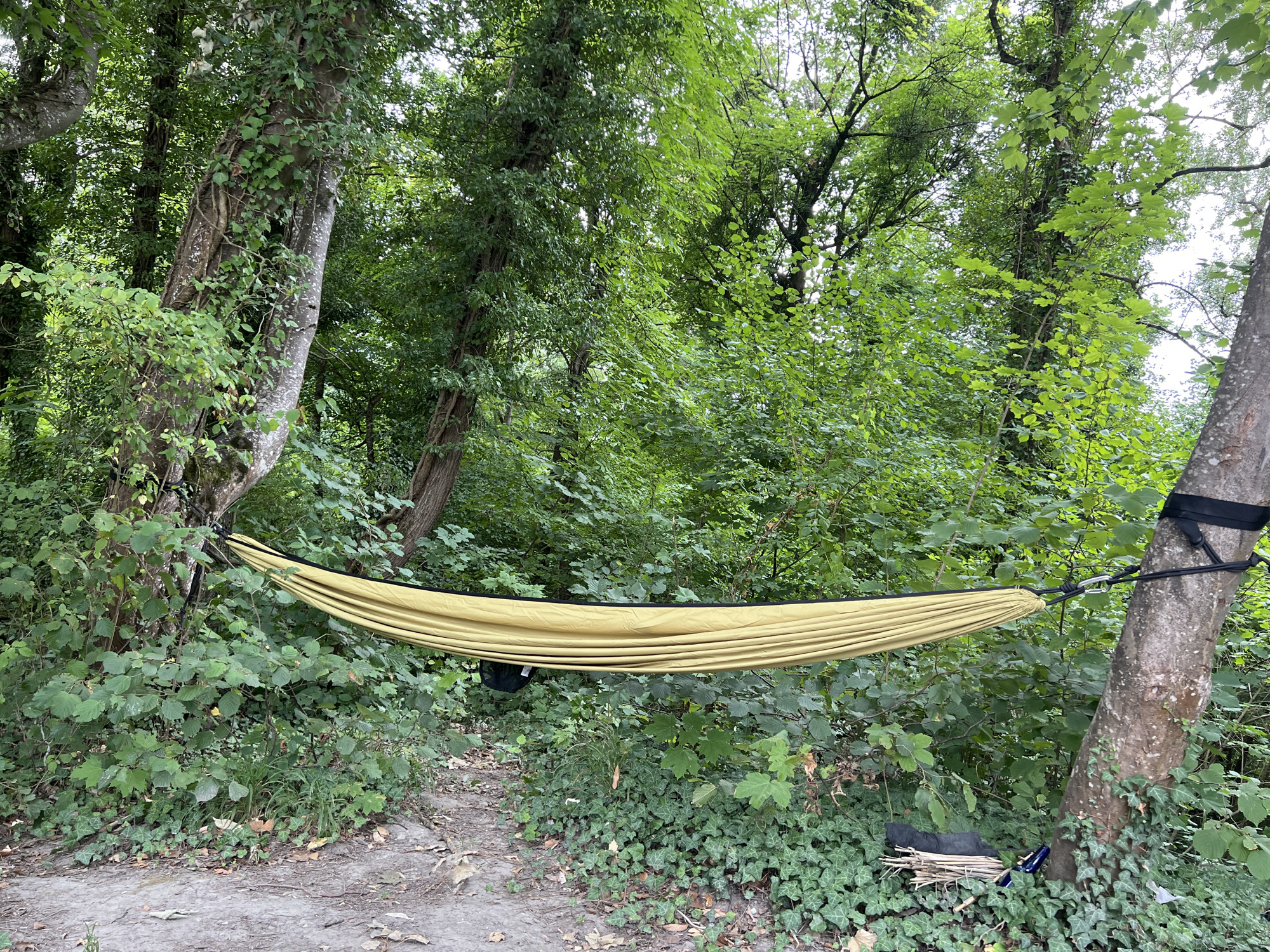 A yellow hammock is suspended between two trees in a wooded area. The trees are tall and covered in green leaves, and the ground is covered in dirt and fallen leaves. The hammock is empty and appears to be made of nylon. There is a black bag hanging from the hammock. The scene is peaceful and suggests a place for relaxation and nature enjoyment. The location is Étrembières, France.