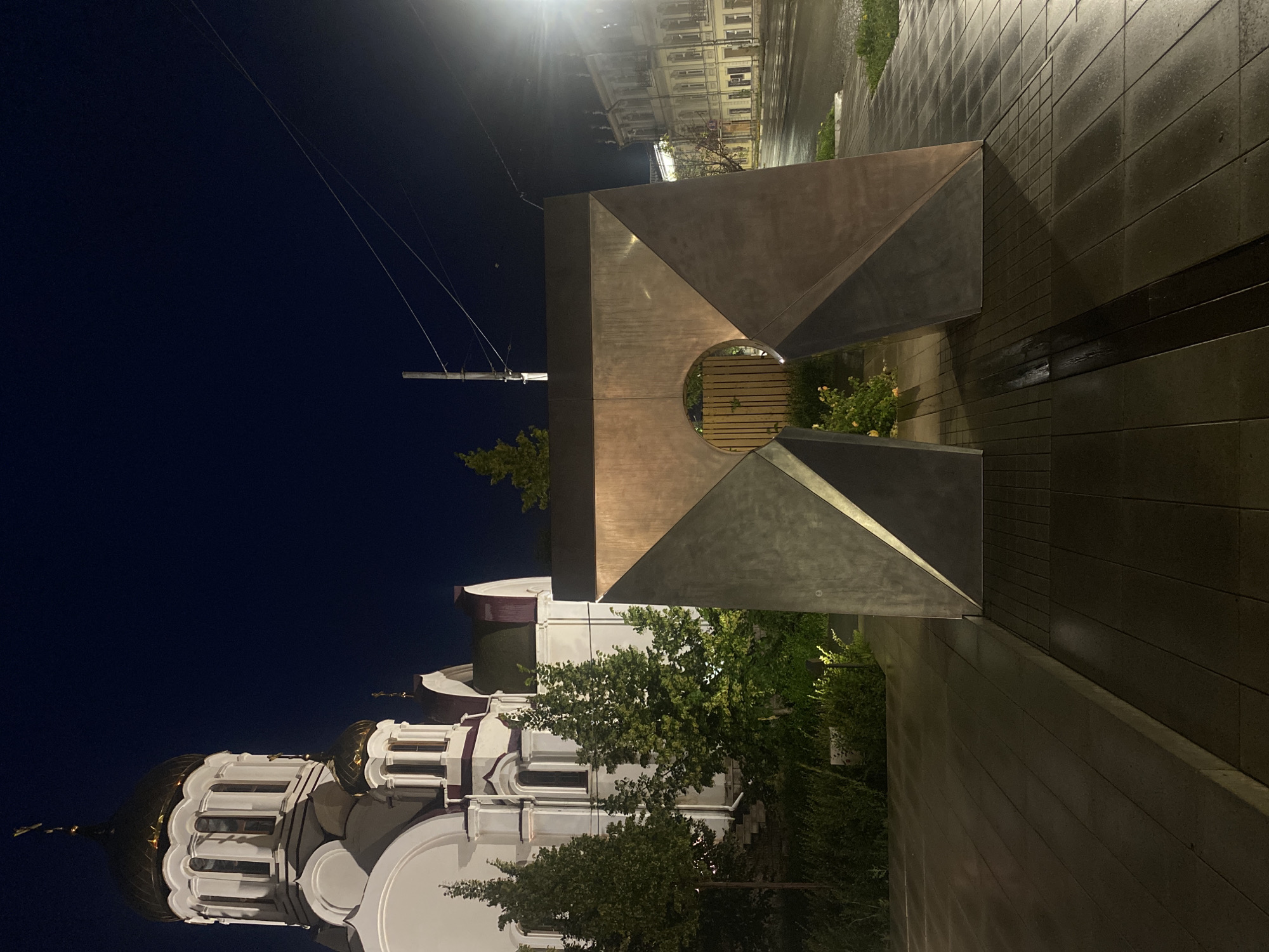 An outdoor, eye-level, full shot captures a modern, abstract sculpture in the foreground, positioned on a paved area. The sculpture is composed of angular, metallic planes that form an archway with a circular opening. Behind the sculpture, a white church with golden domes and red accents is visible, partially obscured by lush green trees. To the right of the sculpture, a street runs alongside an old, multi-story building with a facade of windows and architectural details, illuminated by streetlights. The sky is a deep, dark blue, indicating nighttime. The ground is wet and reflective, suggesting recent rain. The scene is devoid of any people.