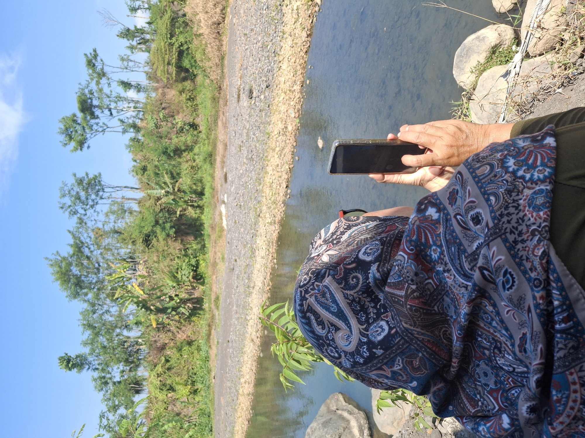 A person wearing a headscarf with a paisley pattern is standing by a river, holding up a smartphone to take a picture. The person is facing away from the camera, with their right hand holding the phone. The river has clear, shallow water with a rocky bed. The riverbank is lined with stones and vegetation, and beyond that, there are trees and a grassy embankment under a clear blue sky. The scene appears to be in a rural or natural area, possibly in Lumajang, Indonesia, given the vegetation and terrain.