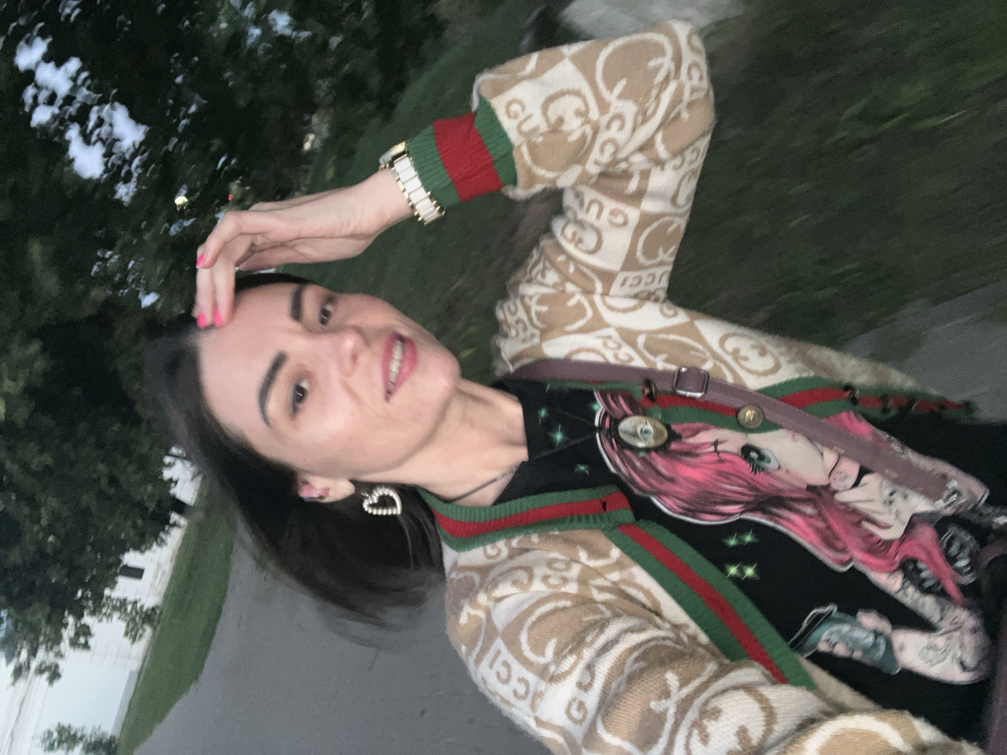 The image shows a woman taking a selfie outdoors in Krasnoe-na-Volge, Russia. She is smiling and touching her hair with her right hand, while her left arm is extended towards the camera. She is wearing a beige cardigan with a repeating "GUCCI" logo pattern, and a black t-shirt with a graphic print of a pink-haired anime-style character. The cardigan has red and green striped cuffs and collar. She is also wearing a silver watch on her left wrist and a heart-shaped earring in her left ear. A strap from a bag is visible across her chest. The background consists of trees and grass, suggesting it is daytime. The lighting indicates it might be dusk or overcast.