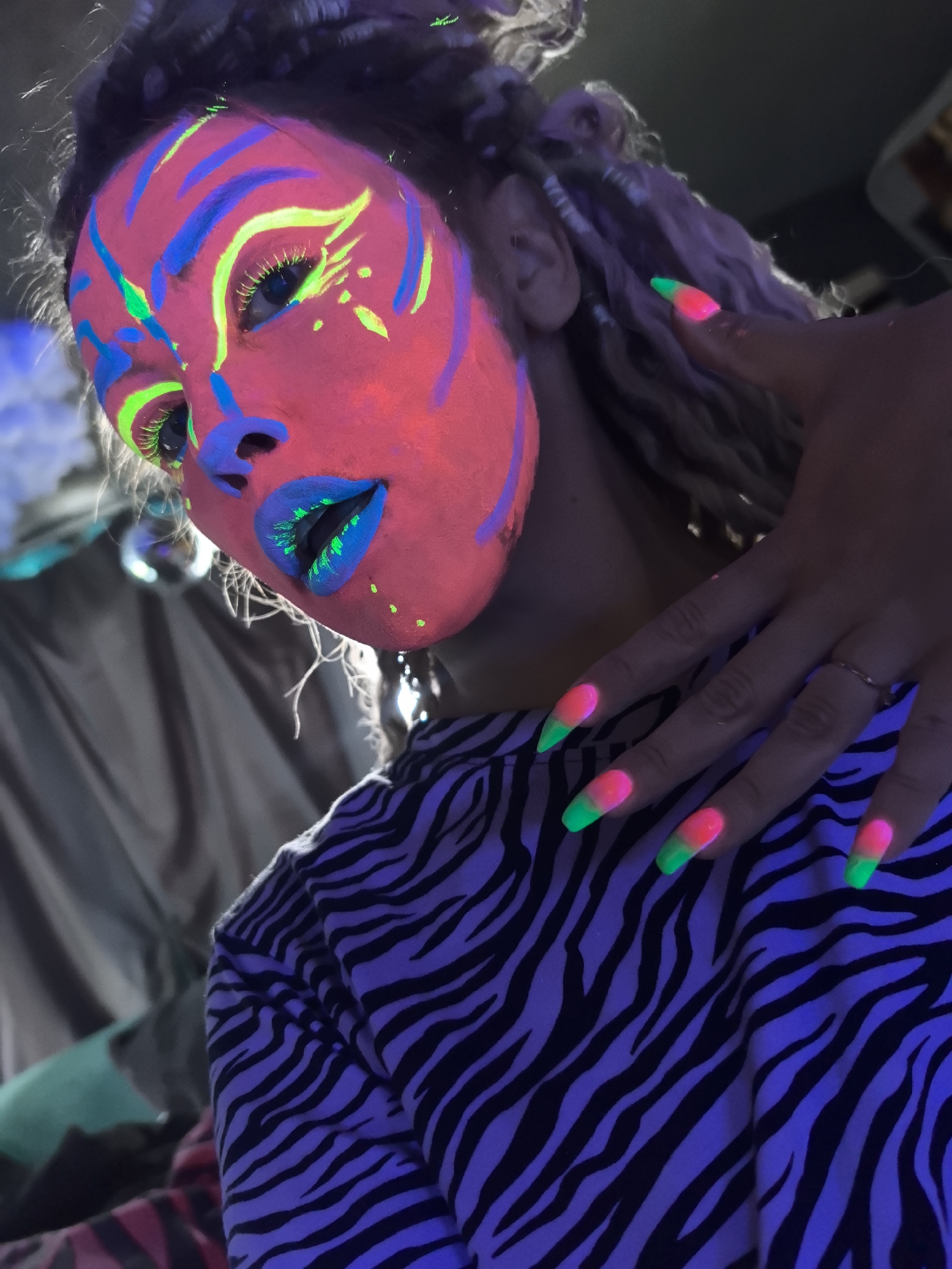 A person with light skin is depicted in a close-up shot, showcasing vibrant neon makeup. The face features a pink base adorned with blue and neon green accents, including stripes and eye makeup. The lips are colored blue, with neon green highlights. The person's hair appears to be styled in light-colored braids. The hand displays long nails painted with a gradient of pink and neon green. The person is wearing a black and white zebra-striped shirt. The background is partially visible, with a mix of shadowy objects. The image is stylized and appears to be shot under blacklight or UV lighting, resulting in the glowing effect. There is no information to indicate this image was taken in Noviy, Russia.