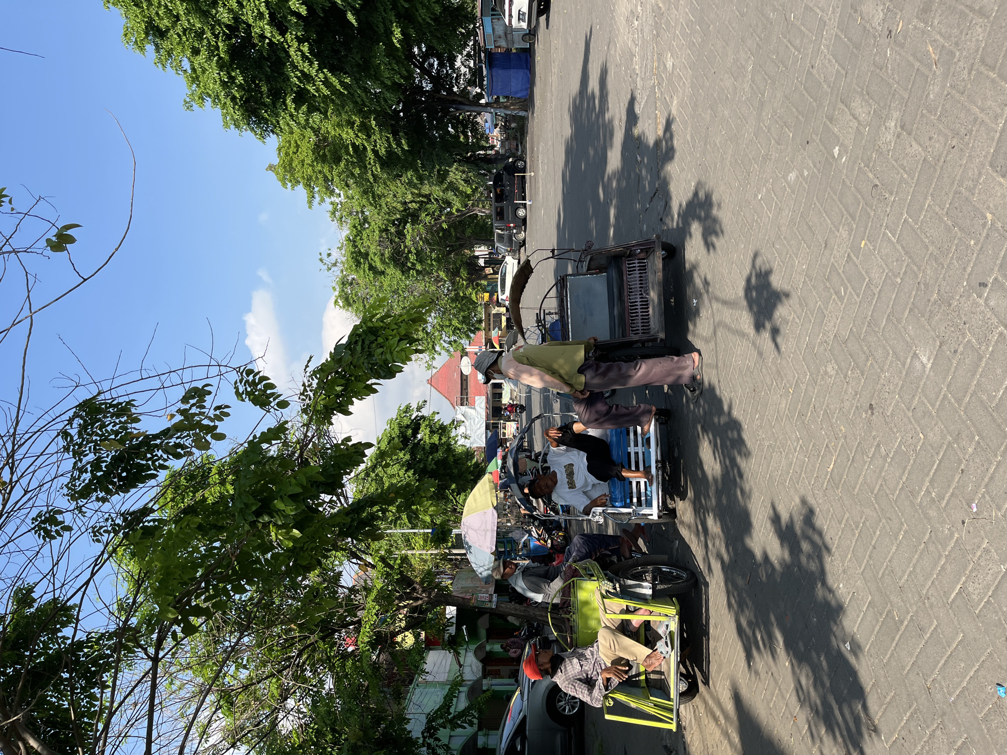 In Juwet Kenongo, Indonesia, on a bright, sunny day, several people are gathered near three-wheeled vehicles that appear to be rickshaws or modified motorcycles. The scene is set in an outdoor area with a paved ground surface, likely a street or market square, lined with trees providing dappled shade.
In the foreground, a man wearing a red cap and a plaid shirt sits in a bright yellow rickshaw. To his right, another man in a cap and a gray jacket is seated in a white and blue rickshaw, facing the viewer. A third rickshaw, darker in color, is positioned further back.
Several individuals are standing and interacting, seemingly engaged in conversation or waiting for passengers. One man, wearing a light green shirt and dark pants, stands near the second rickshaw. Another man, in a white shirt and dark pants, is seated in the same rickshaw, with his legs extended.
In the background, there are buildings with red roofs, and a variety of vehicles, including cars and what appears to be a black van. Various stalls or temporary structures, one covered with a blue tarp, are also visible, suggesting a marketplace or transport hub. The overall atmosphere is relaxed and casual, characteristic of daily life in a local Indonesian setting.