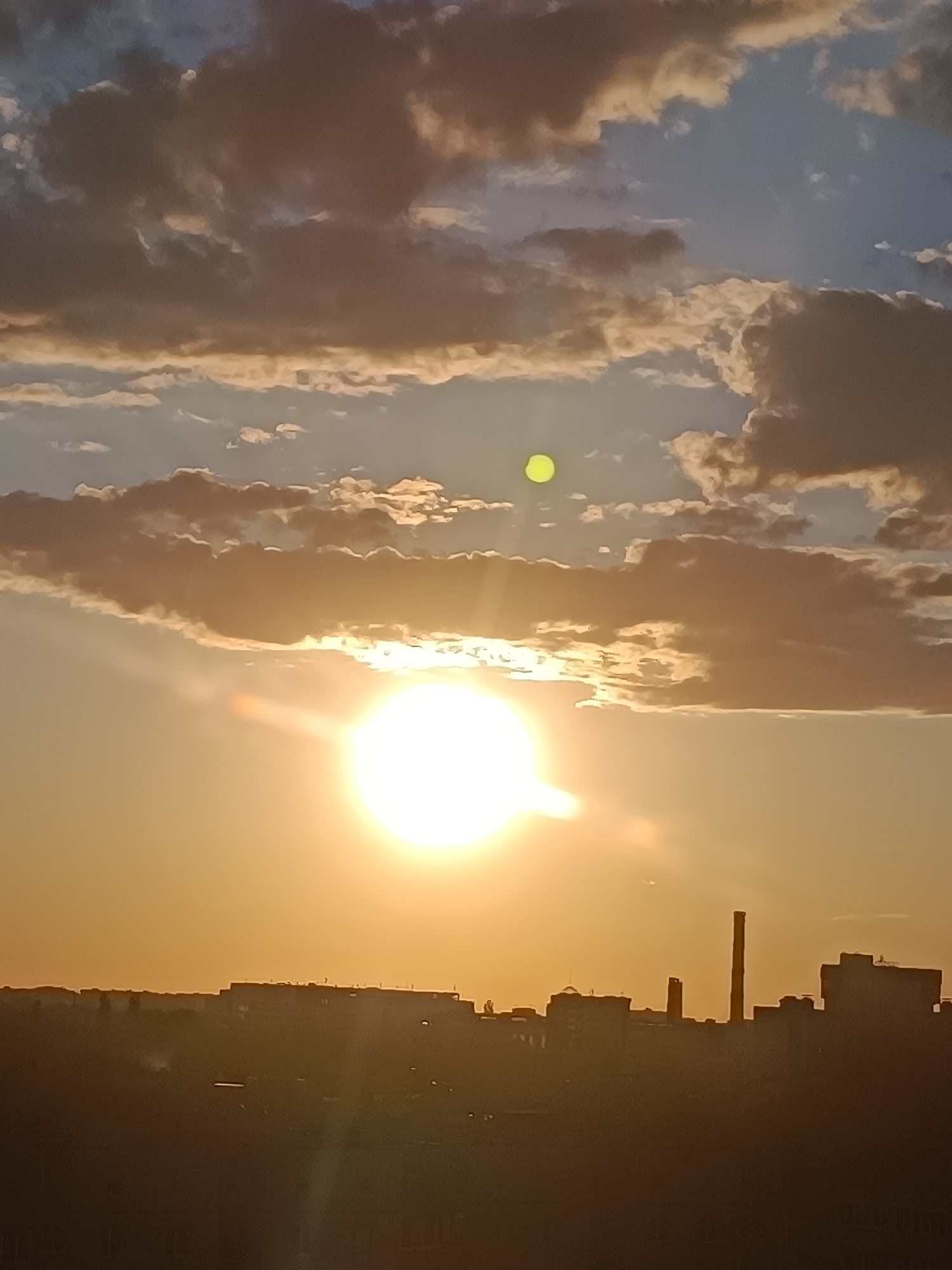 The image captures a sunset over the city of Brest, Belarus. A large, bright sun is positioned in the lower center of the frame, casting a warm, orange glow across the sky and illuminating the clouds. The clouds are a mix of dark gray and lighter, golden hues, suggesting a dramatic atmospheric effect. A faint green orb, possibly lens flare or a secondary atmospheric phenomenon, is visible in the upper left quadrant of the sky.
In the foreground, a silhouetted cityscape extends across the horizon. Buildings of various heights are visible, including a prominent tall, slender smokestack, indicative of an industrial area. The overall impression is one of an industrial city bathed in the beautiful, fading light of sunset. There are no visible people or animals in the frame. The scene is static and peaceful, characterized by the interplay of light, cloud formations, and the urban silhouette.