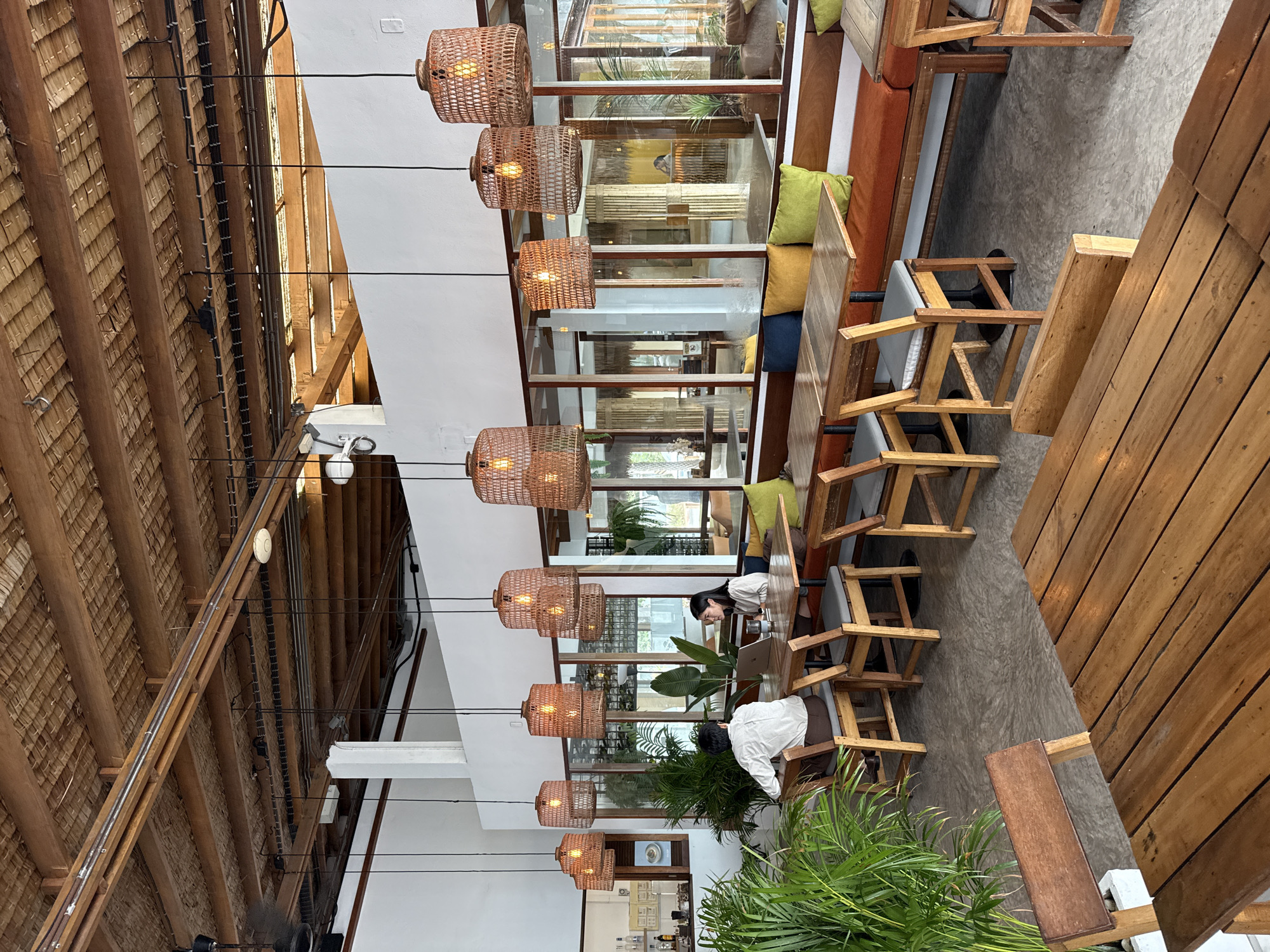 This image depicts the interior of a restaurant or cafe, likely located in Ko Samui District, Thailand, characterized by its tropical ambiance. The ceiling features exposed wooden beams and a thatched roof structure, with a visible ventilation fan on the left. Numerous woven pendant lights, made of natural fibers, hang from the ceiling, illuminating the space with a warm glow.
In the mid-ground, two individuals are seated at a wooden table. A person wearing a white shirt is facing away from the camera, while another person, wearing a light-colored top, is looking towards them. A laptop and drinks are present on their table. The seating area includes long, wooden benches with colorful cushions in shades of orange, yellow, and green. Large potted plants, including a prominent palm, add to the tropical feel.
The flooring appears to be a smooth, light-colored concrete. Through large glass windows, reflections reveal more of the restaurant's interior and possibly an outdoor area. The overall aesthetic suggests a relaxed, natural, and contemporary dining environment.