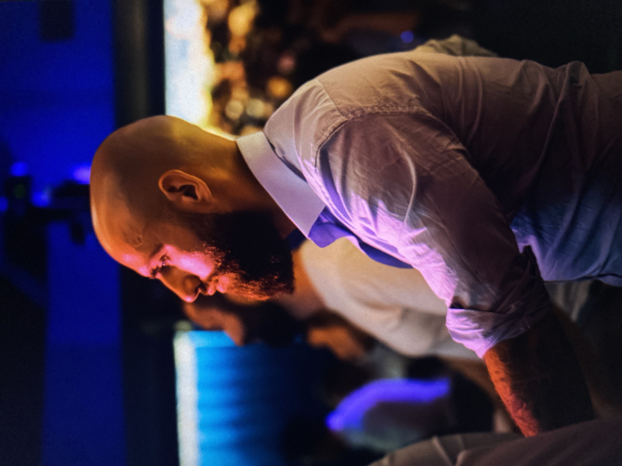 The image portrays a bald man with a beard, seen in profile, intently focused as he leans forward. He is wearing a light-colored button-down shirt, which appears light blue or white in ambient light but takes on a purple-pink hue from the surrounding illumination. His bald head, face, and beard are brightly lit by a strong, warm, reddish-orange light, creating prominent highlights and contours. His right arm, partially visible beneath a rolled-up sleeve, also reflects this warm light.
The background is largely dark and blurred, creating a shallow depth of field that emphasizes the man. Vibrant, out-of-focus light sources populate the background: a dominant blue light emanates from the left, while warmer, bokeh-like orange and yellow lights are visible further back and to the right. This dramatic, colored lighting suggests an indoor setting, likely a stage, studio, or technical area, characteristic of a nighttime event or a controlled environment.
The man's posture and intense downward gaze suggest deep concentration, possibly on an object, screen, or equipment directly in front of him. His expression appears serious or contemplative, fully absorbed in the activity. There are faint, indistinct shapes in the blurred background, hinting at other figures or elements within the scene. No text is visible in the image. The event takes place in Bankya, Bulgaria.